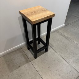 FK TECH - Drewniany taboret barowy z blatem z jasnego drewna i czarną metalową ramą, stojący na szarych płytkach podłogowych przy białej ścianie.