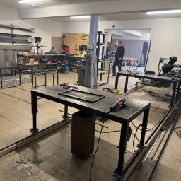 FK TECH - Warsztat spawalniczy z dwoma stołami roboczymi, na jednym leży rama metalowa, a obok szlifierka kątowa, w tle pracownik w czarnym ubraniu.