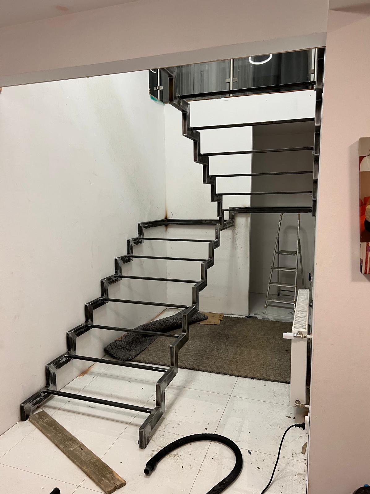 Metalowa konstrukcja schodów w trakcie montażu, widoczne spawy i niedoskonałości wykończenia, drabina w tle, brak poręczy.