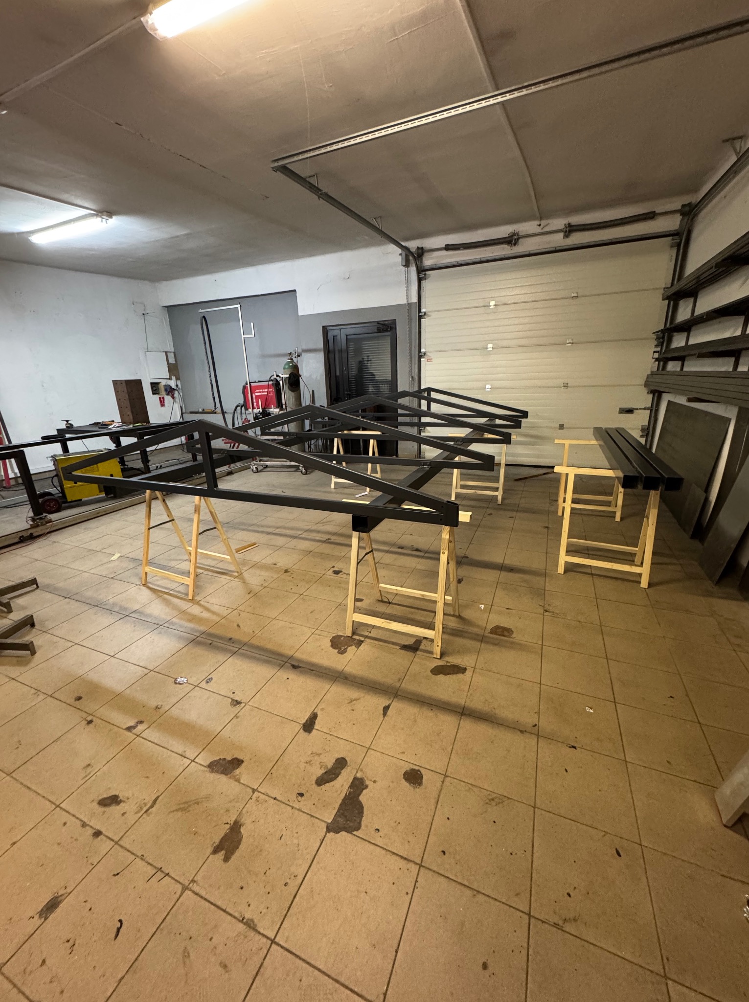 Elementy konstrukcji metalowej ogrodzenia pomalowane na czarno, ustawione na drewnianych kozłach w warsztacie, widoczny sprzęt spawalniczy i brama garażowa.