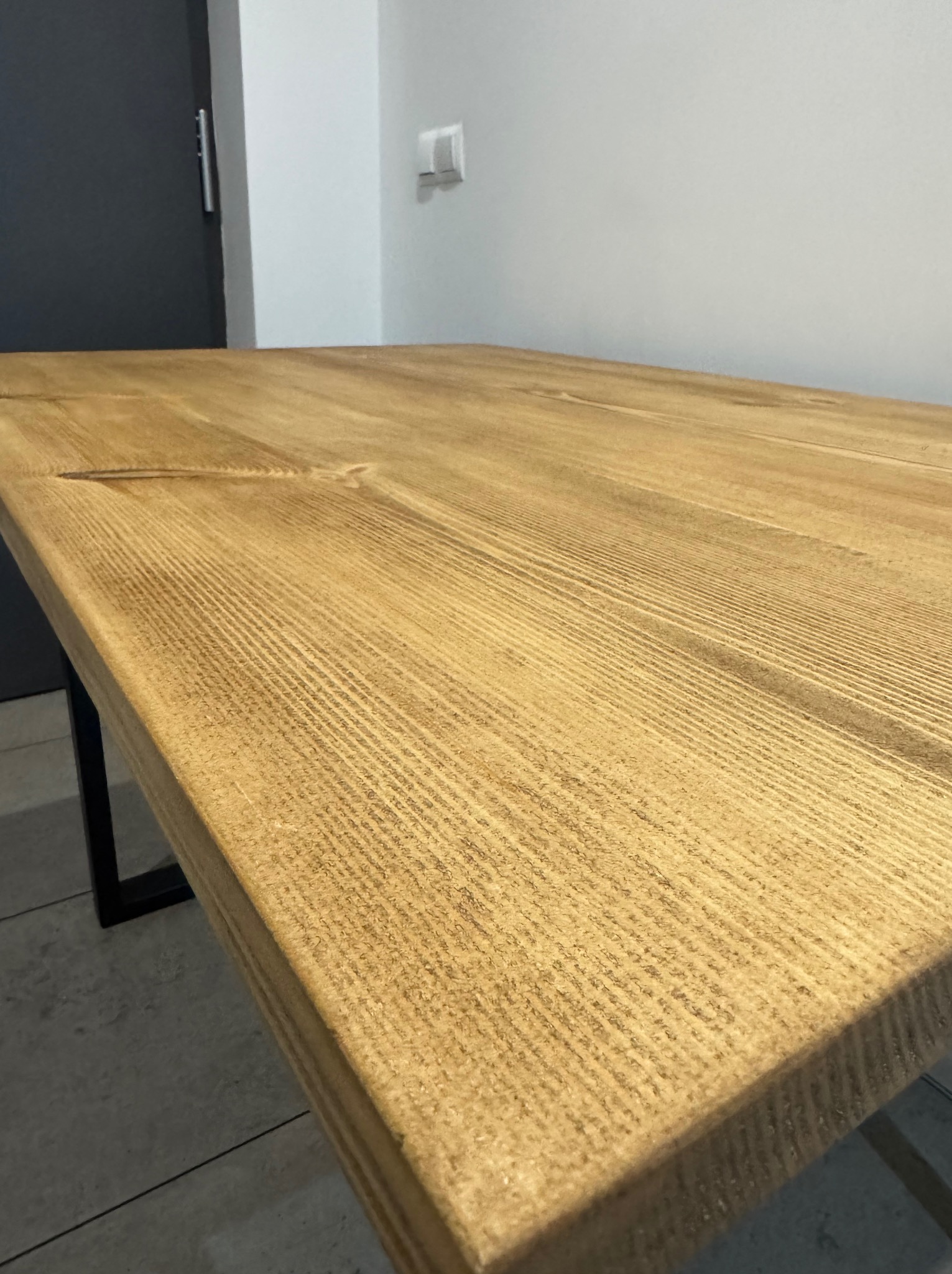 Blat stołu z jasnego drewna o wyraźnym usłojeniu, wsparty na metalowych nogach, widoczny pod kątem z bliska.