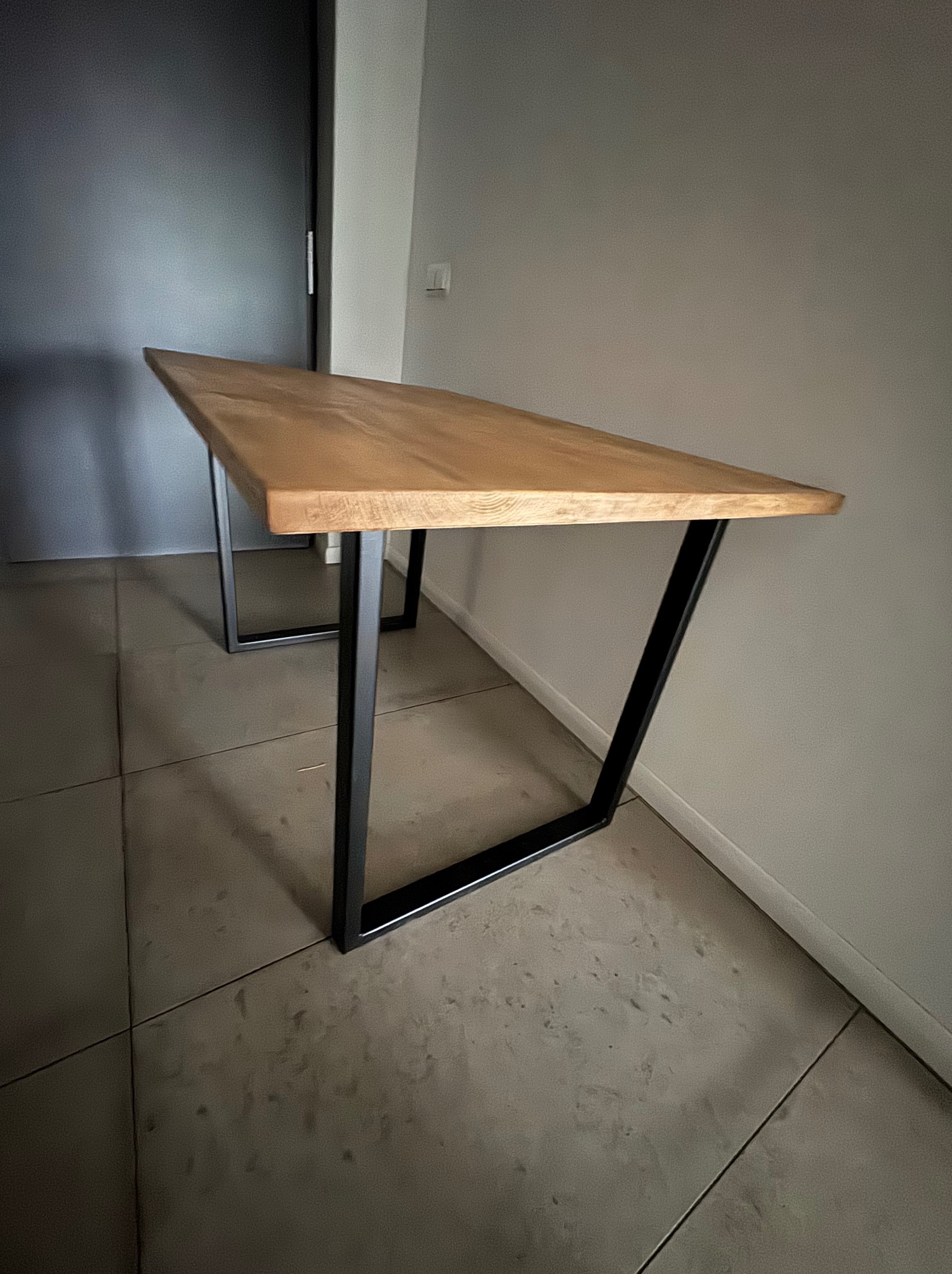 Stół z blatem z jasnego drewna i czarnymi, metalowymi nogami w kształcie trapezu, ustawiony na szarej podłodze z płytek.