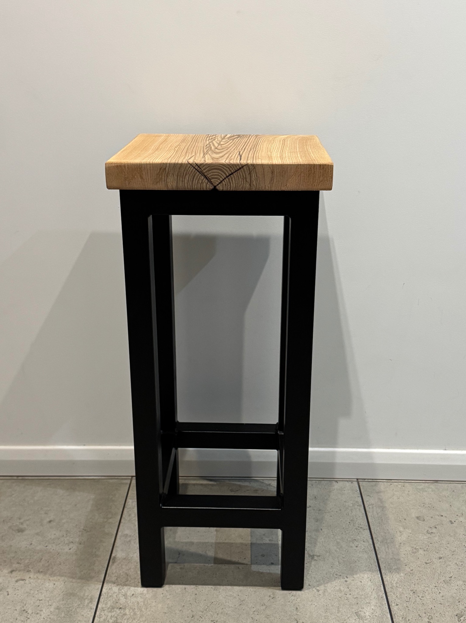 Drewniany taboret z blatem z jasnego drewna o widocznym usłojeniu i czarną, metalową ramą na tle białej ściany i szarej podłogi.