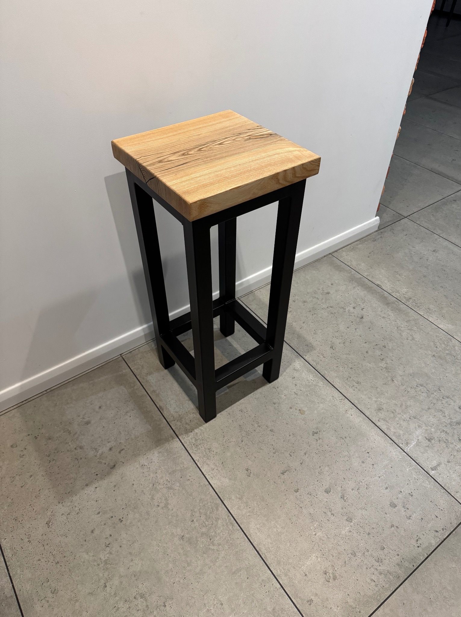 Drewniany taboret barowy z blatem z jasnego drewna i czarną metalową ramą, stojący na szarych płytkach podłogowych przy białej ścianie.