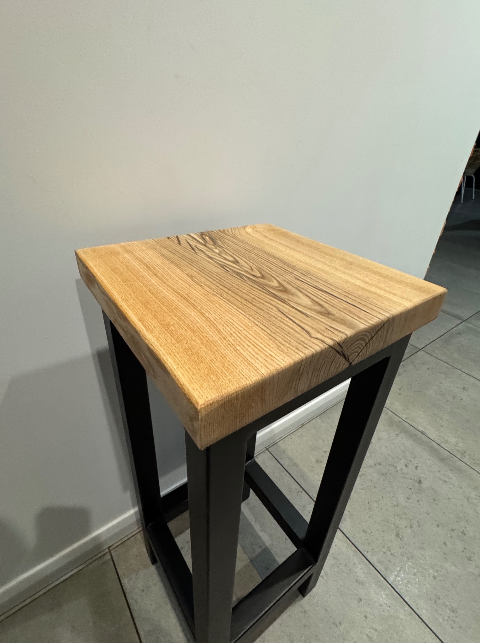 Nowoczesny stołek barowy z blatem z jasnego drewna o wyraźnym usłojeniu i czarną, metalową podstawą w kształcie trapezu, stojący na szarej podłodze przy jasnej ścianie.