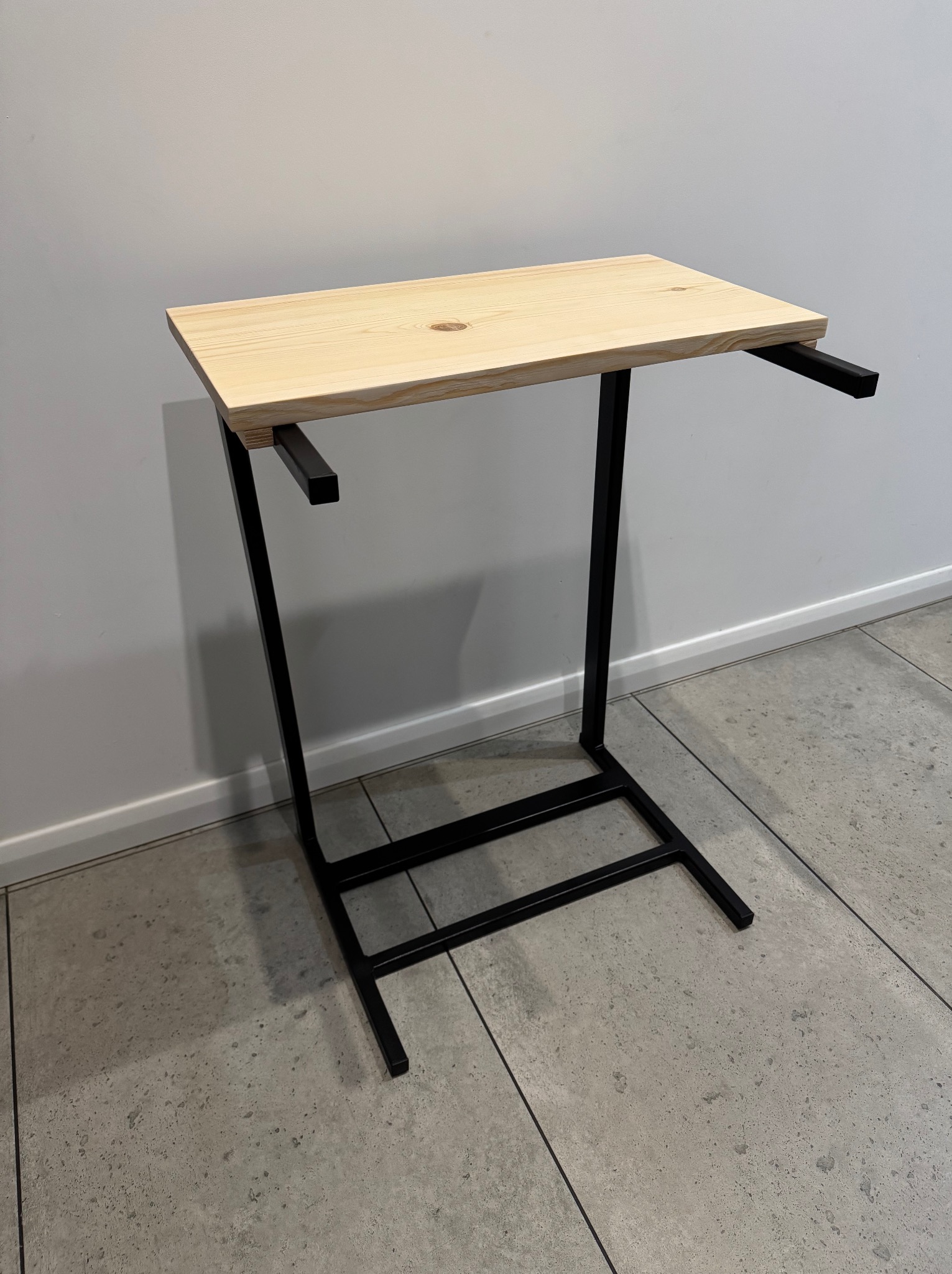 Stolik pomocniczy z jasnego drewna z czarną, metalową podstawą w kształcie litery Z, na tle szarej ściany i podłogi z płytek.