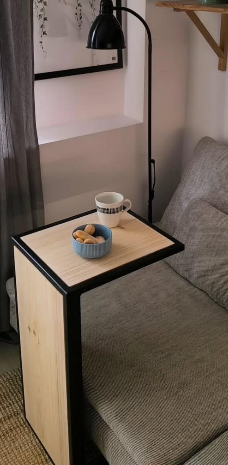 Stolik pomocniczy z jasnego drewna z czarną ramą, ustawiony obok sofy, na blacie niebieska miseczka z ciasteczkami i kubek.