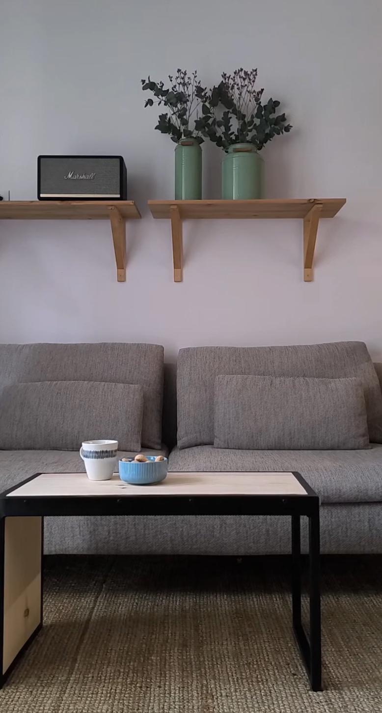 Stolik kawowy z blatem z jasnego drewna i metalową ramą, na nim filiżanka i miseczka, w tle szara sofa, drewniane półki z głośnikiem i wazonami.