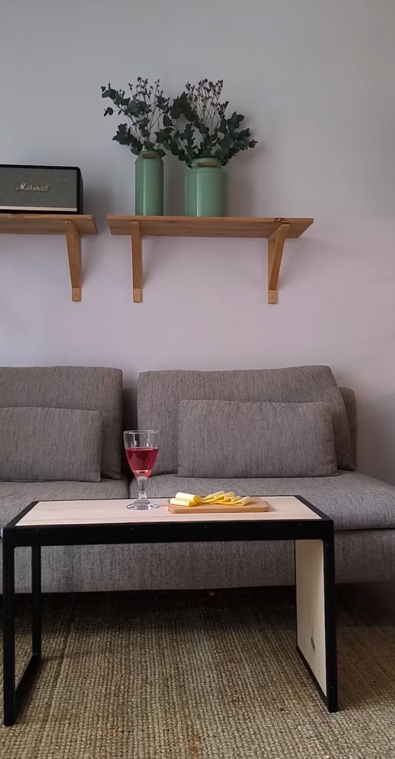 Stolik kawowy z metalowymi nogami i drewnianym blatem, na nim kieliszek różowego wina i deska z serem, w tle szara sofa, drewniane półki z dekoracyjnymi wazonami i głośnik Marshall.