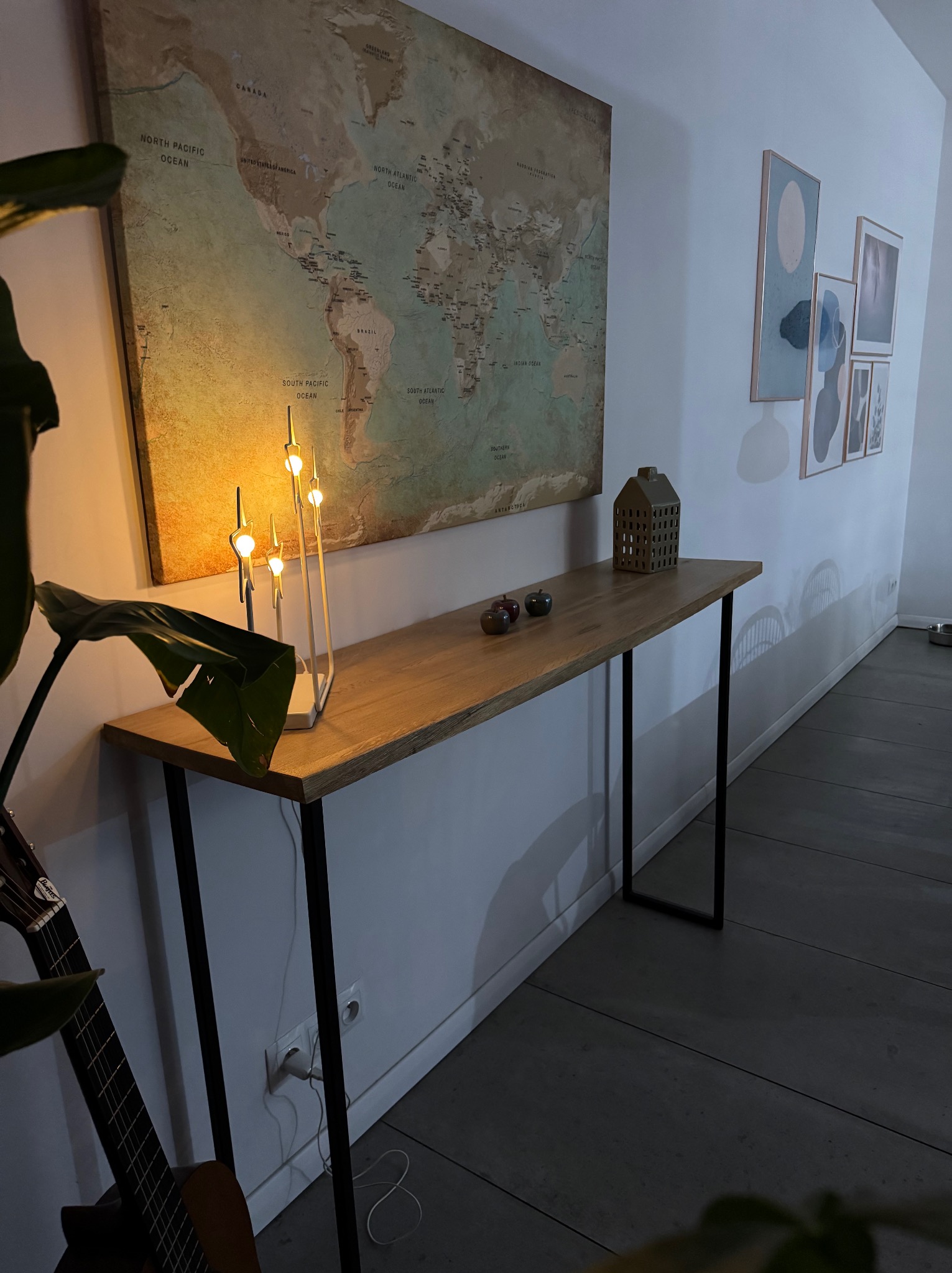 Drewniany blat konsoli na metalowych nogach w minimalistycznym wnętrzu, z dekoracjami: lampką, ceramiczną figurką domu i dwoma jabłkami, na ścianie mapa świata i abstrakcyjne obrazy.
