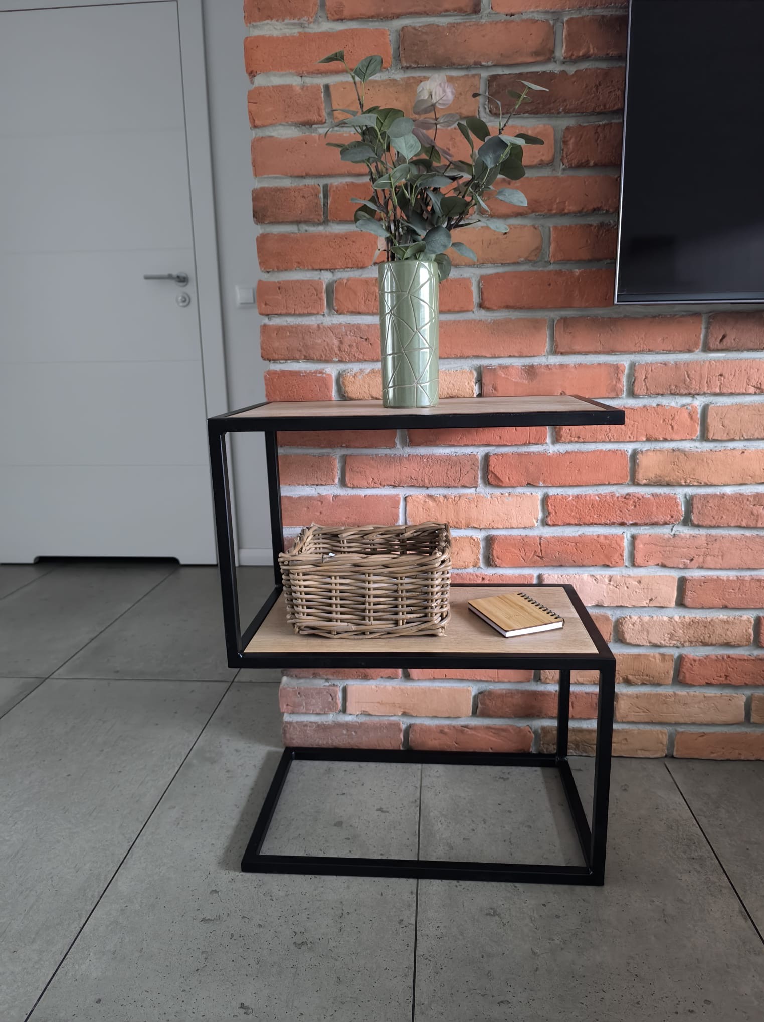 Dwupoziomowy stolik kawowy z czarną metalową ramą i drewnianymi blatami, na tle ceglanej ściany, z wazonem, koszykiem wiklinowym i notatnikiem.