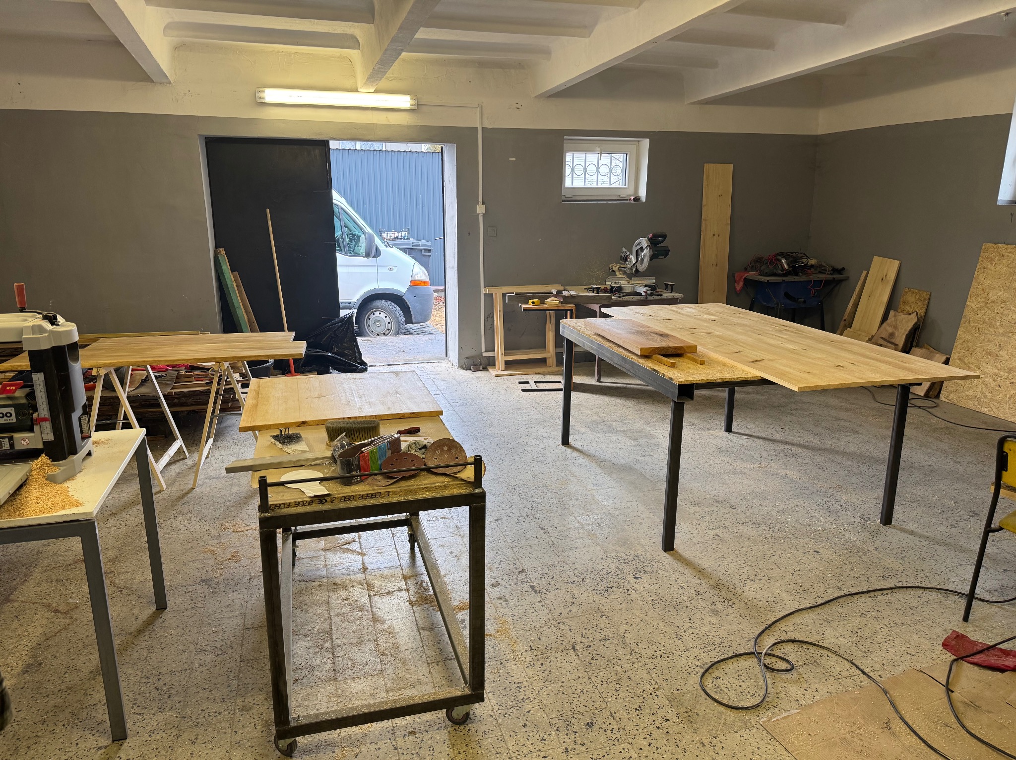 Warsztat z surowymi drewnianymi blatami na metalowych nogach, strugarką, piłą ukośnicą i samochodem dostawczym widocznym w otwartych drzwiach.