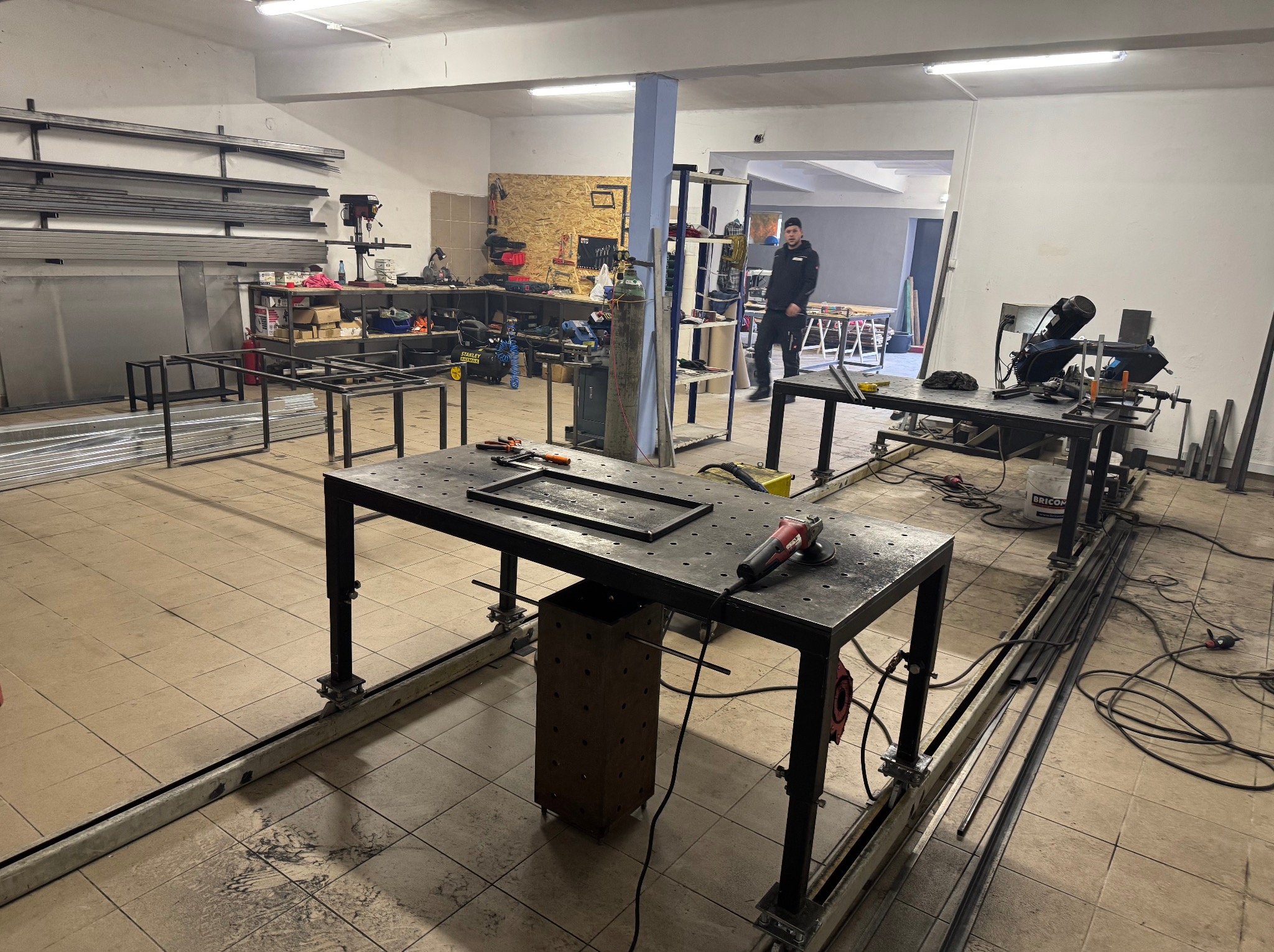 Warsztat spawalniczy z dwoma stołami roboczymi, na jednym leży rama metalowa, a obok szlifierka kątowa, w tle pracownik w czarnym ubraniu.