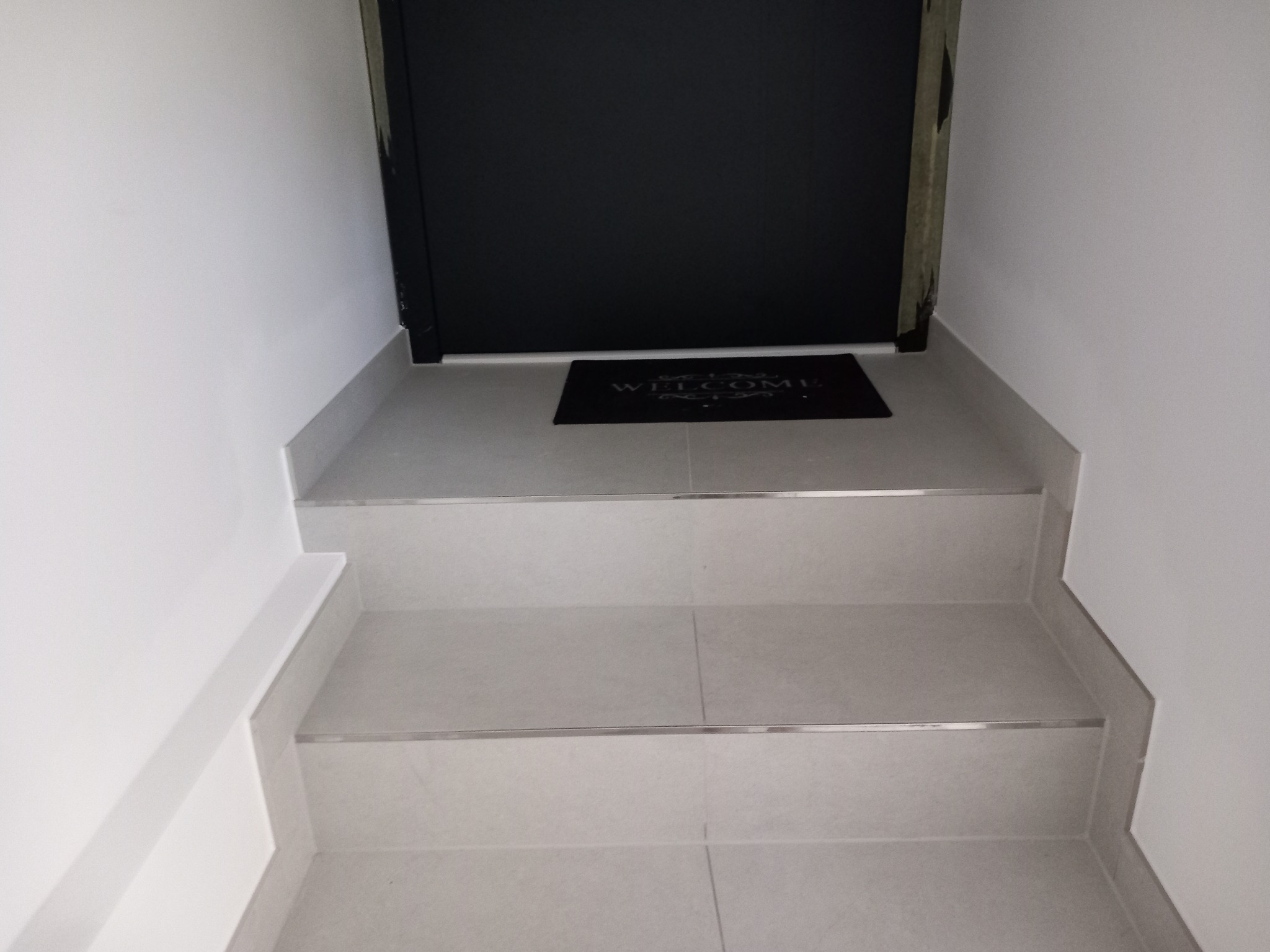 Wykończone schody wejściowe do domu z szarymi płytkami, listwami wykończeniowymi i czarną wycieraczką przed czarnymi drzwiami.