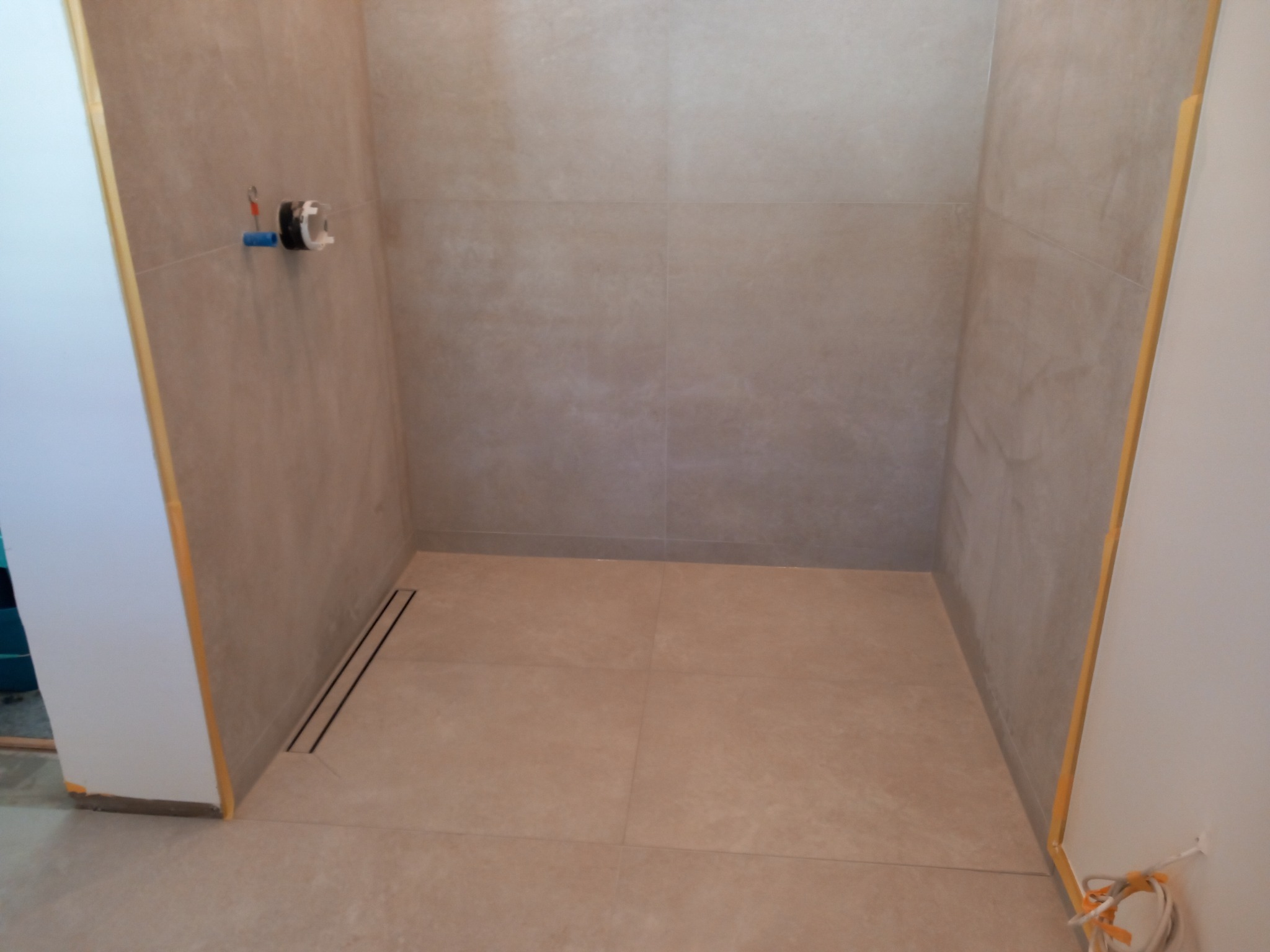 Wnęka prysznicowa wyłożona beżowymi płytkami, z zamontowanym odpływem liniowym i przygotowanymi przyłączami wodno-kanalizacyjnymi na ścianie, widoczne zabezpieczenia narożników.