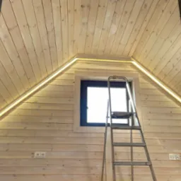 Podświetlenie LED zamontowane w drewnianym wnętrzu poddasza, widoczne gniazdka elektryczne, drabina i okno.