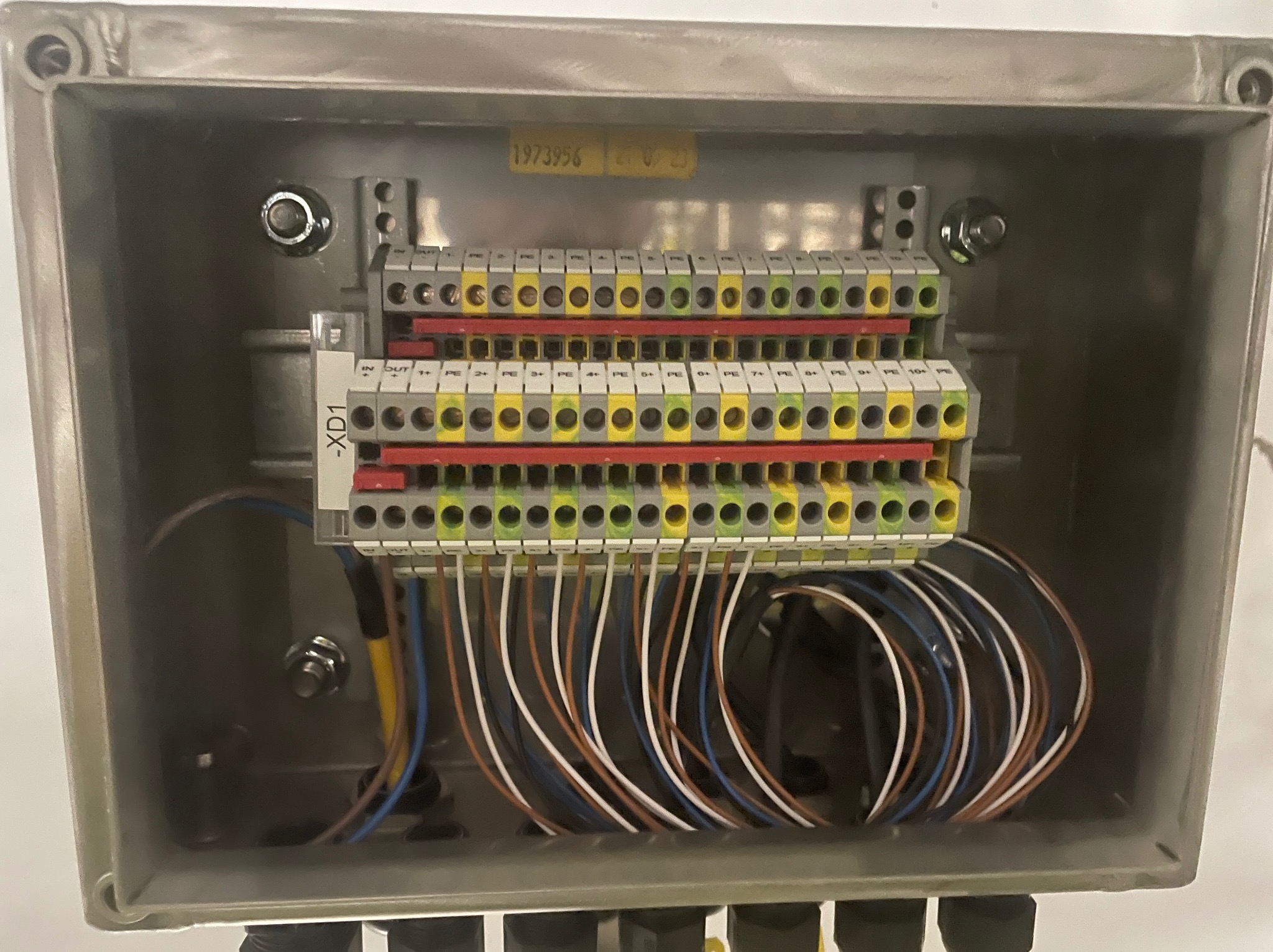 Szara skrzynka elektryczna z listwą zaciskową XD1, do której podłączone są przewody w kolorach brązowym, białym i niebieskim. Widoczna naklejka z numerem seryjnym 1973956 i datą 21.07.23.