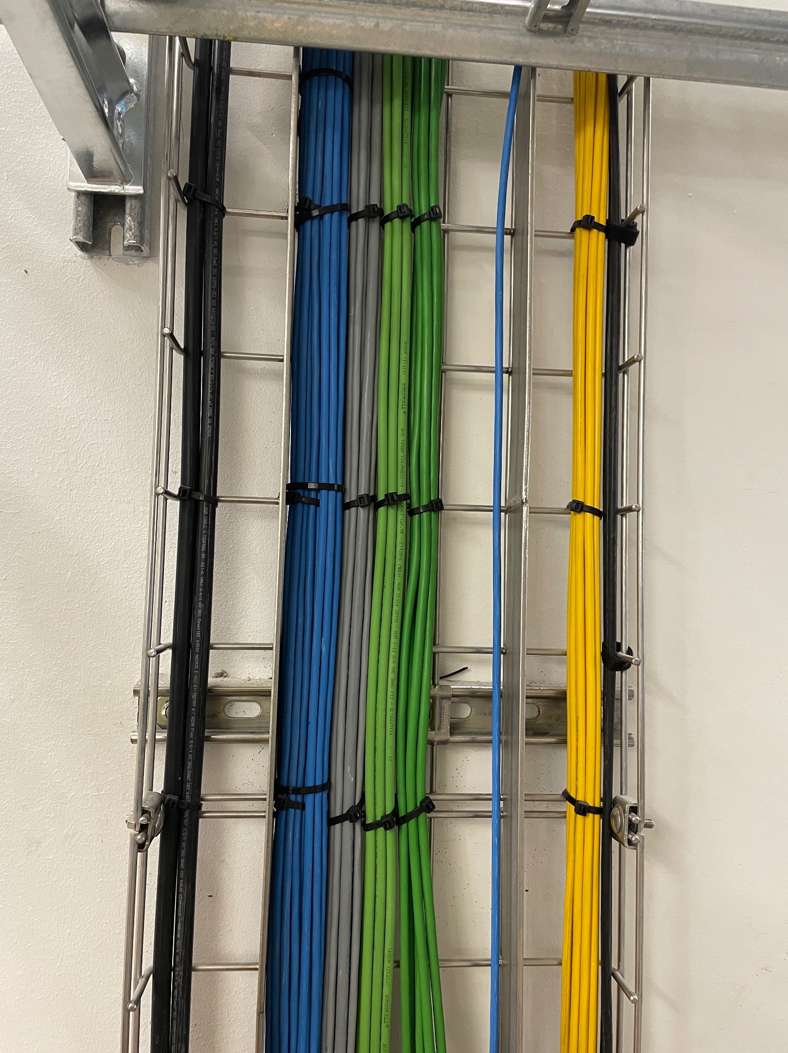 Różnokolorowe przewody elektryczne ułożone pionowo w metalowej korycie kablowej, przymocowane czarnymi opaskami zaciskowymi, widok z bliska na fragment instalacji.