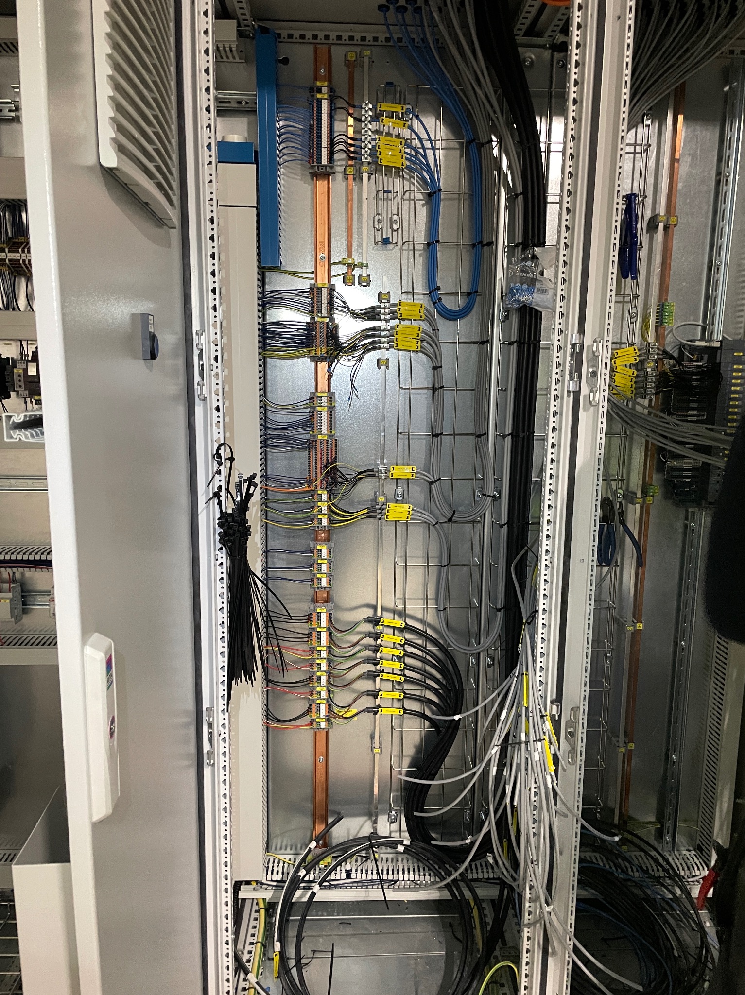 Rozdzielnica elektryczna w trakcie montażu, widoczne uporządkowane przewody, listwy zaciskowe, szyny DIN i metalowa konstrukcja szafy, z wiązką nieużytych opasek kablowych wiszących na boku.