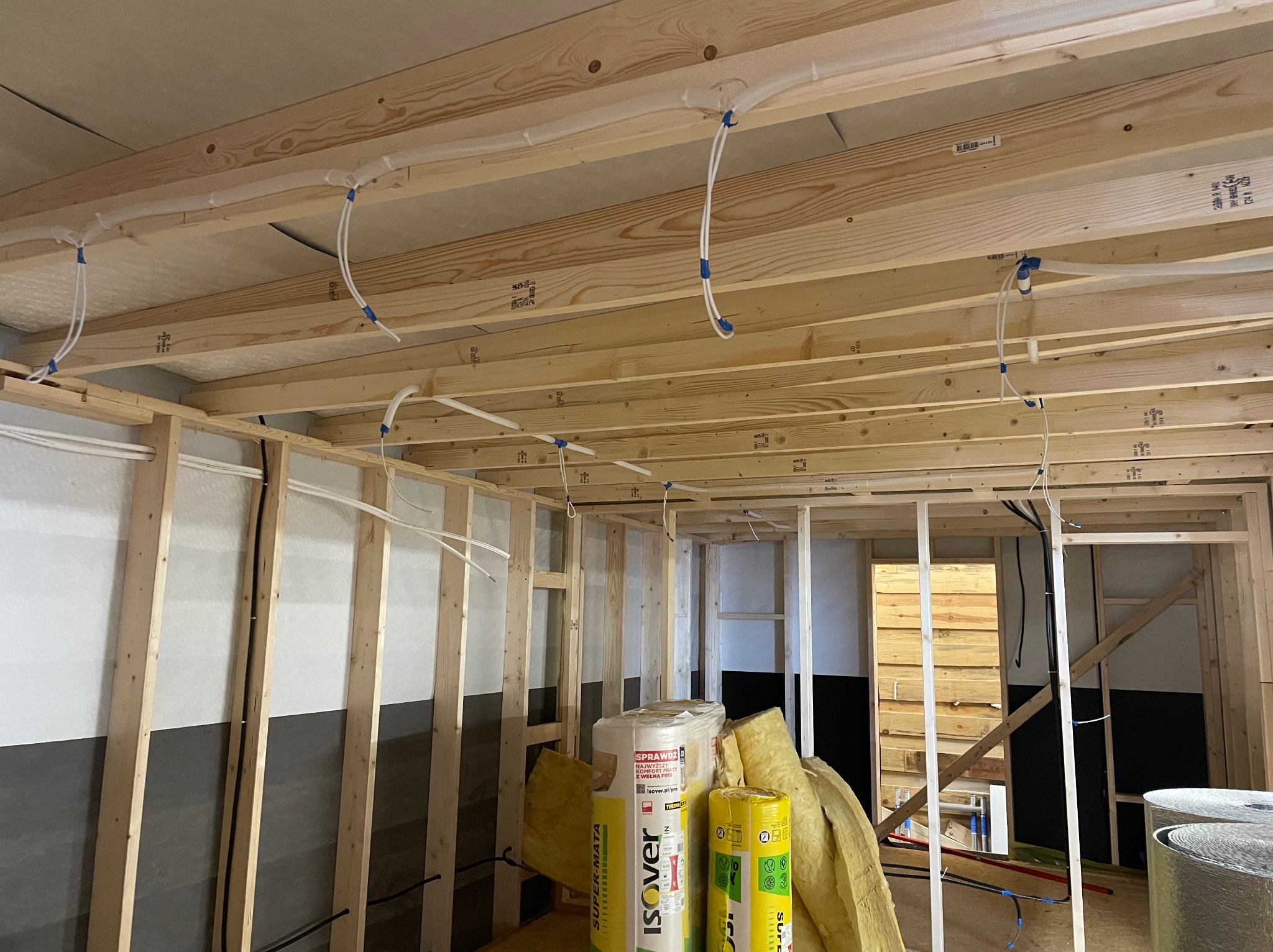 Surowy stan poddasza z widocznymi elementami drewnianej konstrukcji stropu, ociepleniem z wełny mineralnej Isover oraz białymi rurkami instalacji elektrycznej zamocowanymi niebieską taśmą.