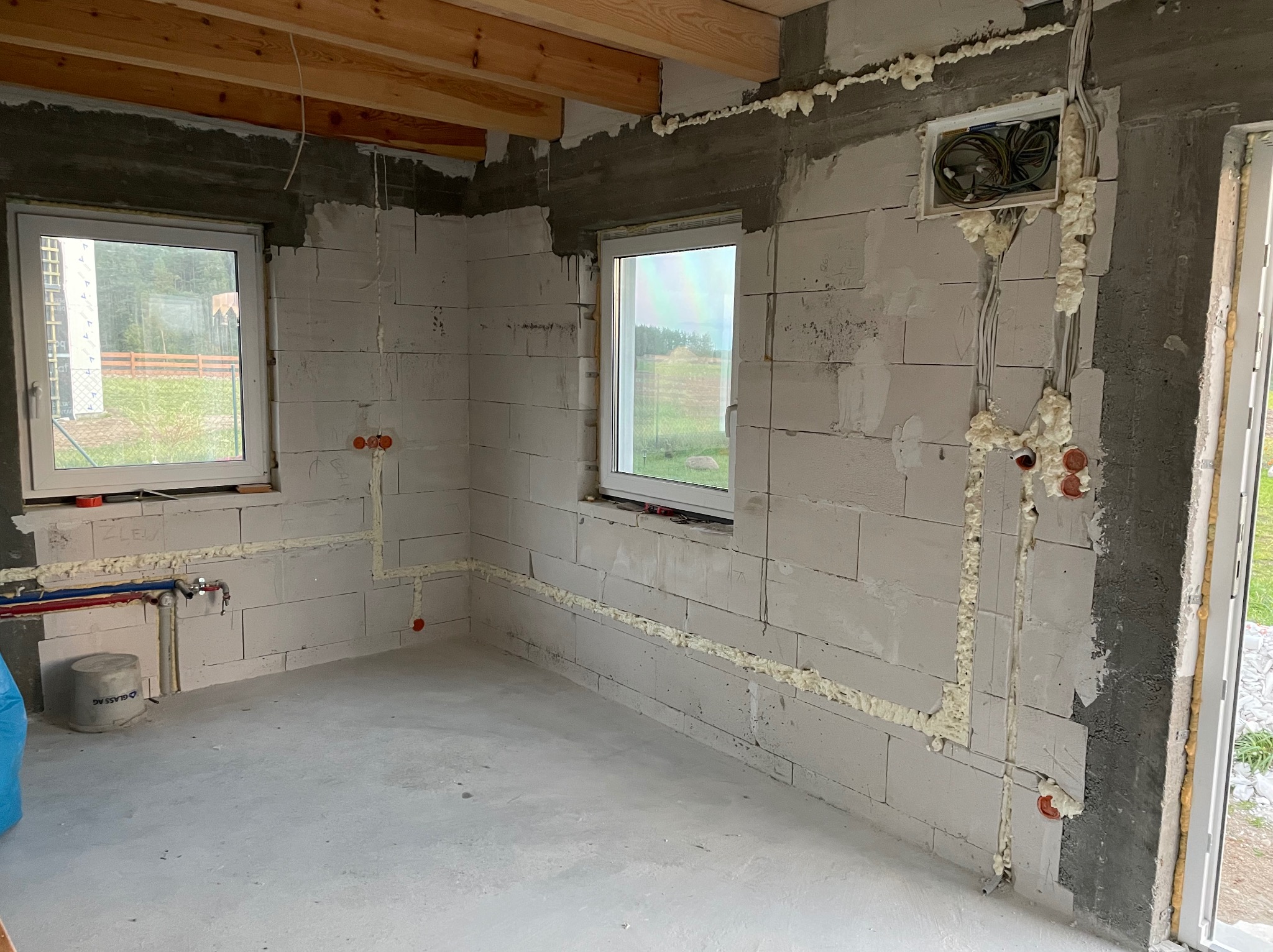 Wnętrze surowego domu w trakcie prac instalacyjnych: widoczne okna, ściany z betonu komórkowego z bruzdami wypełnionymi pianką montażową, wystające kable i rury instalacyjne, surowe drewniane belki...