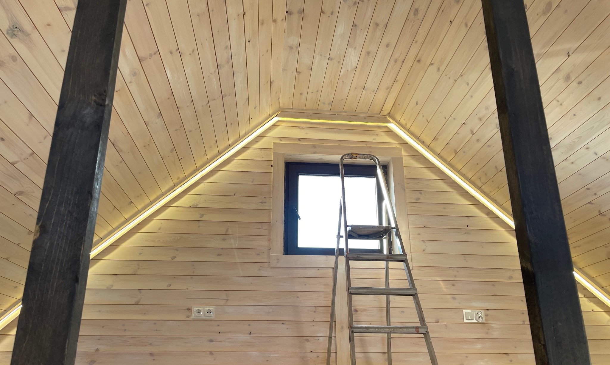 Podświetlenie LED zamontowane w drewnianym wnętrzu poddasza, widoczne gniazdka elektryczne, drabina i okno.