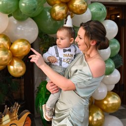 ALINA NIEZGODZIŃSKA - Kobieta w satynowej sukience trzyma rocznego chłopca w ubranku z muchą i mini czapeczce na tle dekoracji z zielonych, złotych i białych balonów oraz kartonowej postaci Simby z Króla Lwa.