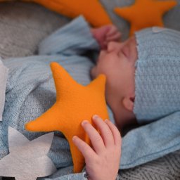 ALINA NIEZGODZIŃSKA - Śpiące niemowlę w błękitnym ubranku i czapce, otoczone pomarańczowymi i białymi gwiazdkami na szarym, dzierganym kocyku.