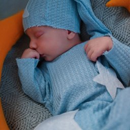 ALINA NIEZGODZIŃSKA - Śpiący noworodek w niebieskim, dzierganym stroju z gwiazdkami, leżący na szarym kocu z pomarańczowymi poduszkami w kształcie księżyca i gwiazd.