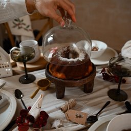 ALINA NIEZGODZIŃSKA - Uroczysty stół z babką pod szklanym kloszem na drewnianym stojaku, ciemne kieliszki, dekoracje świąteczne i ręka zdejmująca klosz.