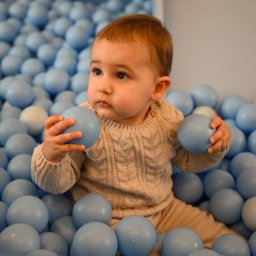 ALINA NIEZGODZIŃSKA - Urocze niemowlę w beżowym sweterku siedzi w basenie wypełnionym błękitnymi piłeczkami, trzymając dwie z nich w rączkach i spoglądając w lewo.