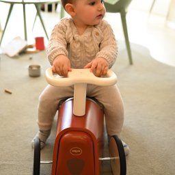 ALINA NIEZGODZIŃSKA - Mały chłopiec w beżowym swetrze i spodniach siedzi na czerwonym jeździku z białym kierownicą w jasnym wnętrzu.