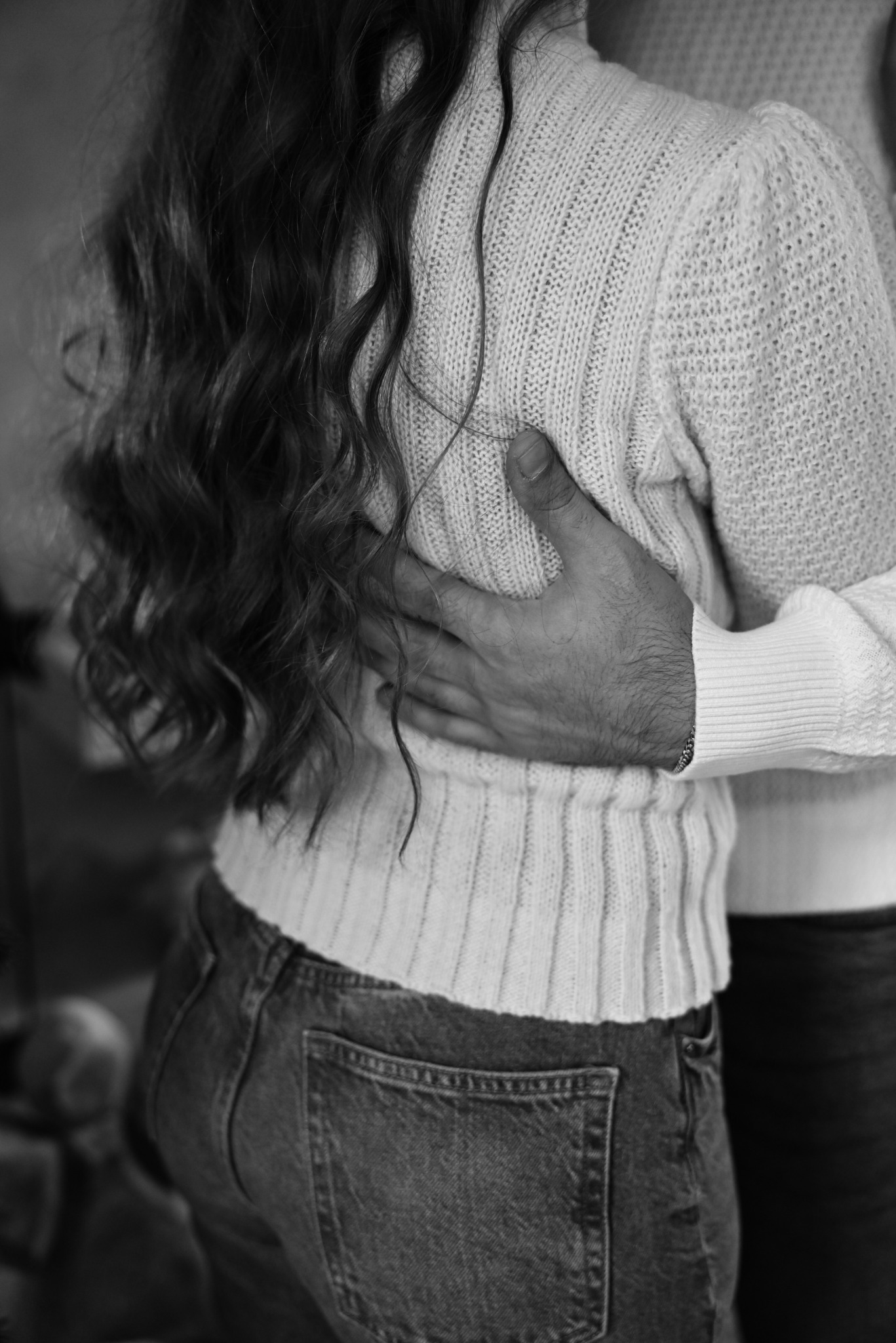 Czarno-białe zbliżenie na parę obejmującą się, widoczne włosy kobiety, dżinsy i sweter, oraz dłoń mężczyzny na jej plecach.