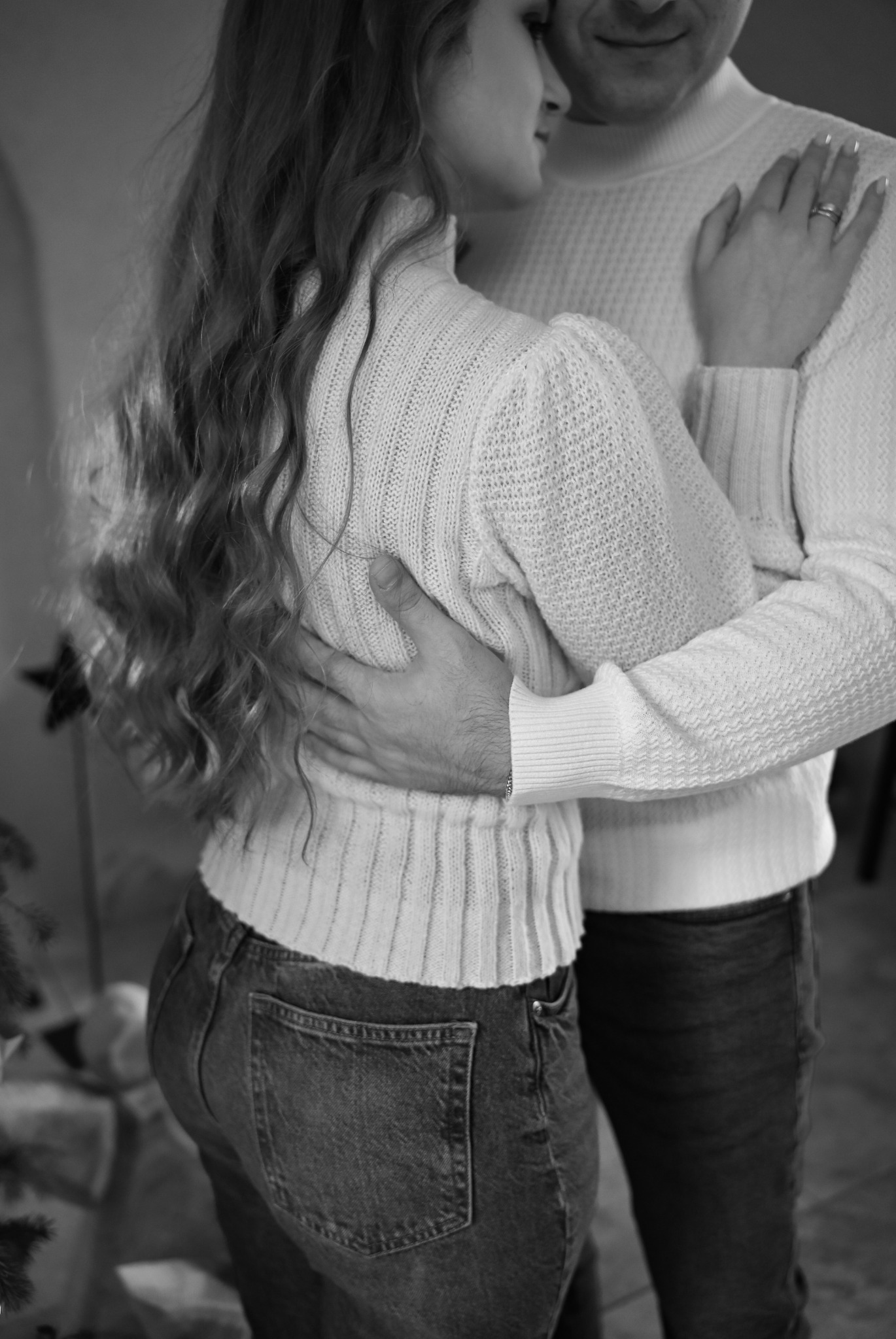 Czarno-białe zdjęcie obejmującej się pary w jasnych swetrach. Kobieta ma długie, falowane włosy i dżinsy, a na palcu mężczyzny widoczny jest pierścionek.