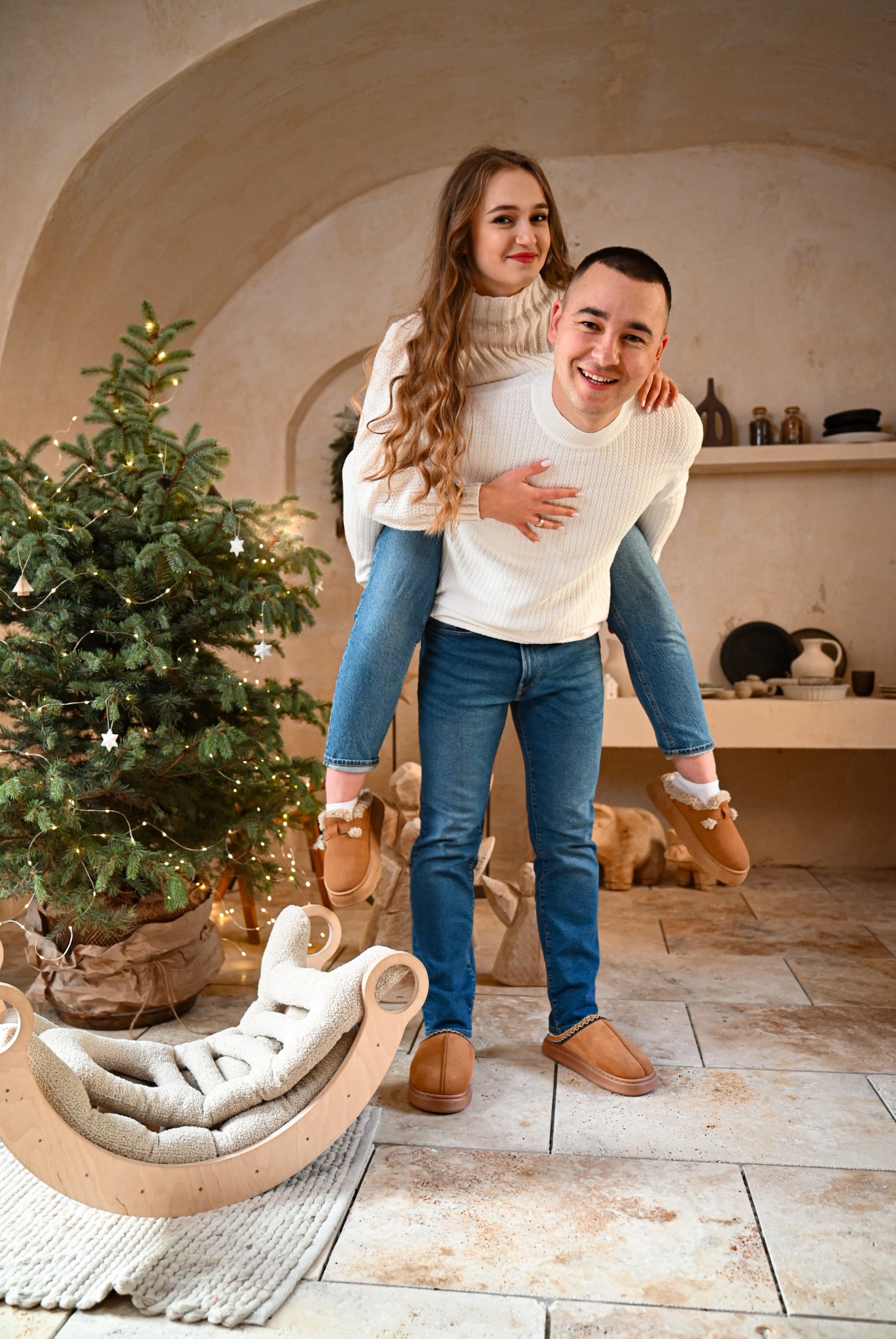 Uśmiechnięta para w jasnych swetrach i jeansach: mężczyzna nosi kobietę na barana przy choince, w tle półki z ceramiką i sufit z łukami, obok bujak dla dziecka.