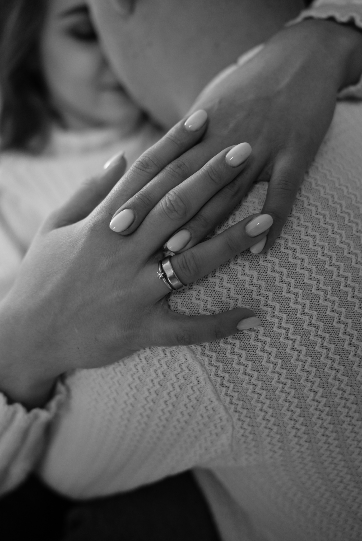 Czarno-białe zbliżenie dłoni z pierścionkiem zaręczynowym spoczywającej na ramieniu drugiej osoby, delikatne paznokcie w odcieniu nude, sweter o wyraźnym wzorze w tle.