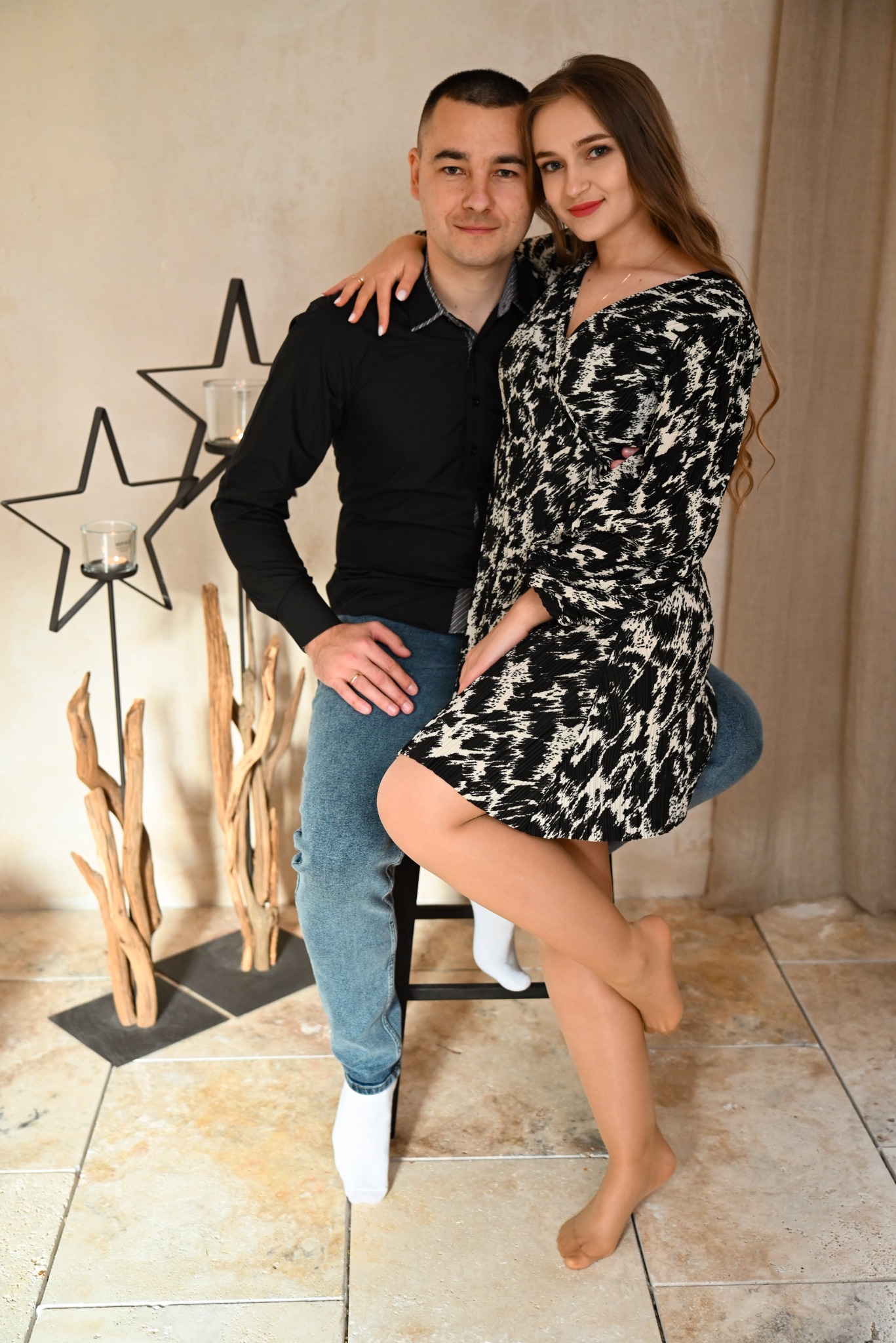 Para: mężczyzna w czarnej koszuli i jeansach siedzi na stołku, obejmując kobietę w czarno-białej sukience, stojącą bokiem z nogą założoną na nogę, w tle dekoracje ze świecznikami w kształcie gwiazd...
