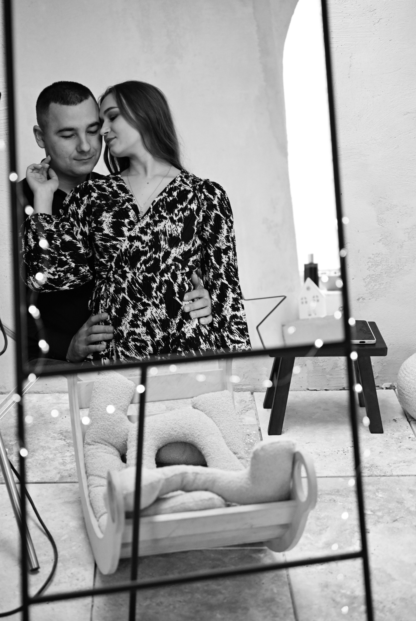 Czułe ujęcie pary w ciąży, widoczne w lustrze, z małym łóżeczkiem dziecięcym w dolnej części kadru i dekoracyjnymi światełkami.