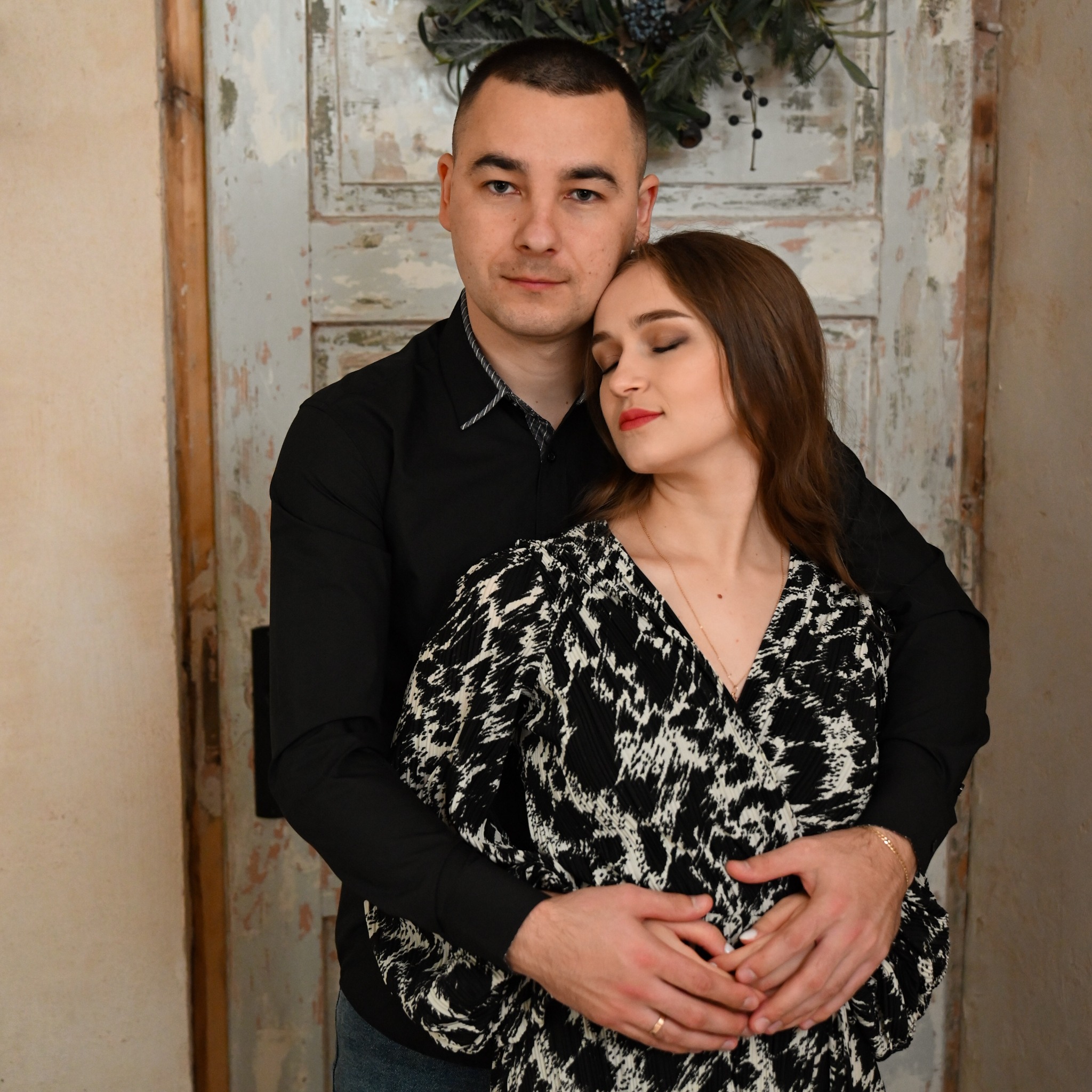 Zakochana para w objęciach na tle starej, malowanej szafy z dekoracją świąteczną, kobieta w wzorzystej sukience z zamkniętymi oczami.