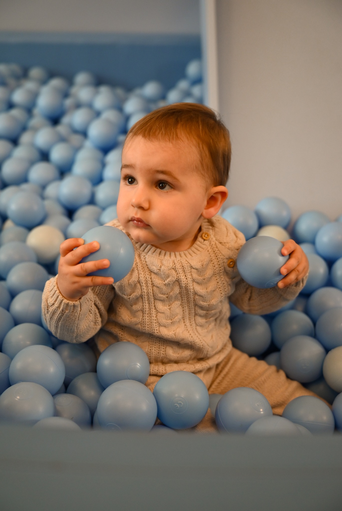 Urocze niemowlę w beżowym sweterku siedzi w basenie wypełnionym błękitnymi piłeczkami, trzymając dwie z nich w rączkach i spoglądając w lewo.