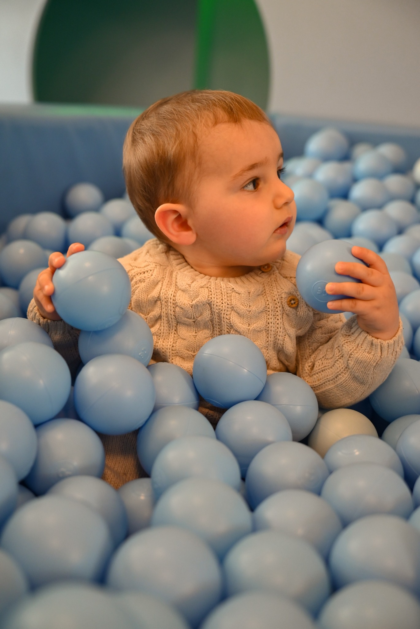Uroczy malec w beżowym sweterku zanurzony w basenie wypełnionym błękitnymi piłeczkami, trzymając po jednej w każdej rączce i z zaciekawieniem spoglądając w bok.
