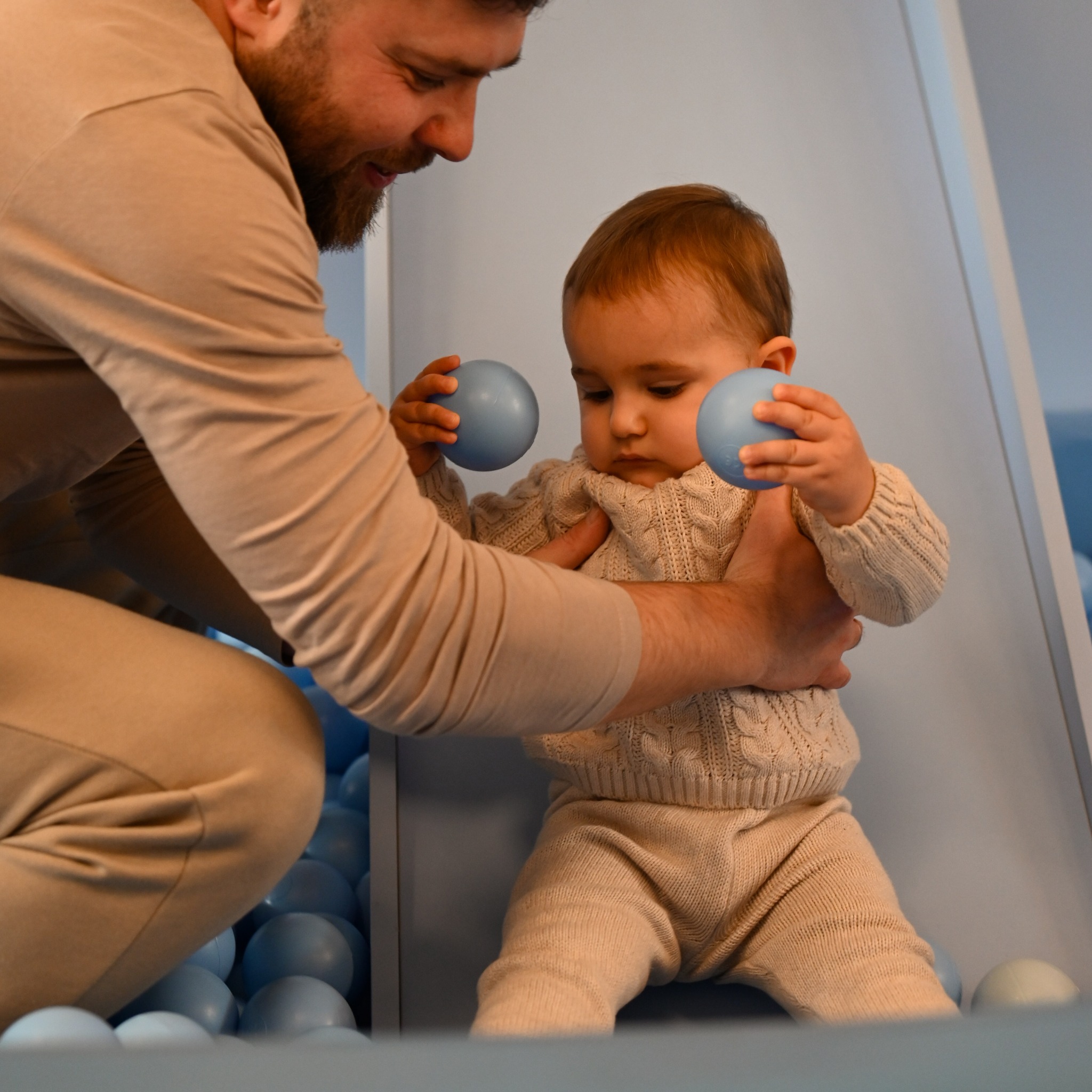 Ojciec wspiera małe dziecko w beżowym swetrze i spodniach, stojące w basenie z błękitnymi piłeczkami, trzymające po jednej piłce w każdej rączce.