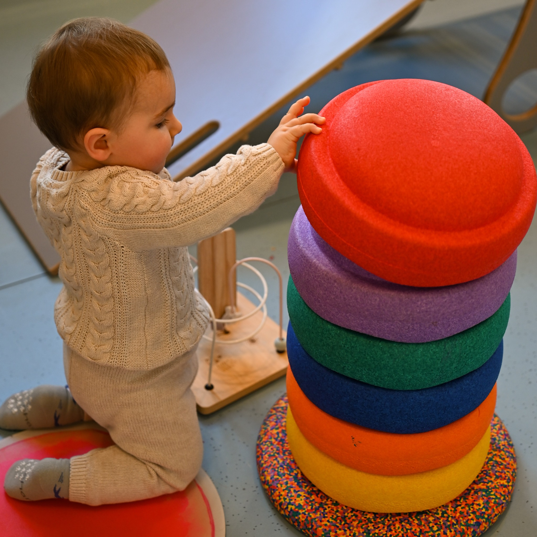 Niemowlę w kremowym swetrze dotyka piramidy z kolorowych, okrągłych poduszek sensorycznych, obok zabawka edukacyjna z pętlami na drewnianej podstawie.