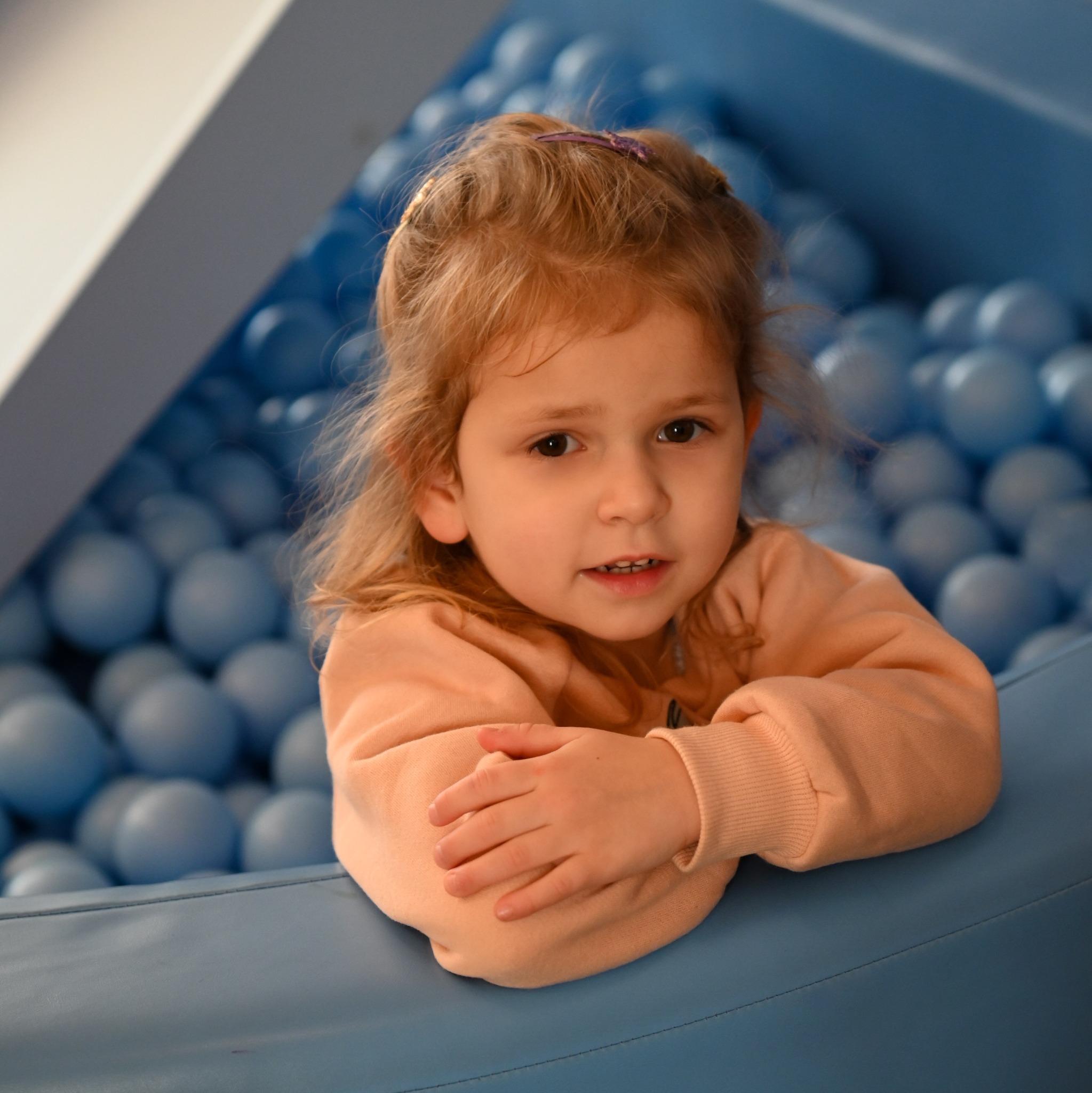 Dziewczynka w brzoskwiniowej bluzie opiera się o brzeg basenu wypełnionego niebieskimi piłeczkami, z fioletową spinką we włosach.