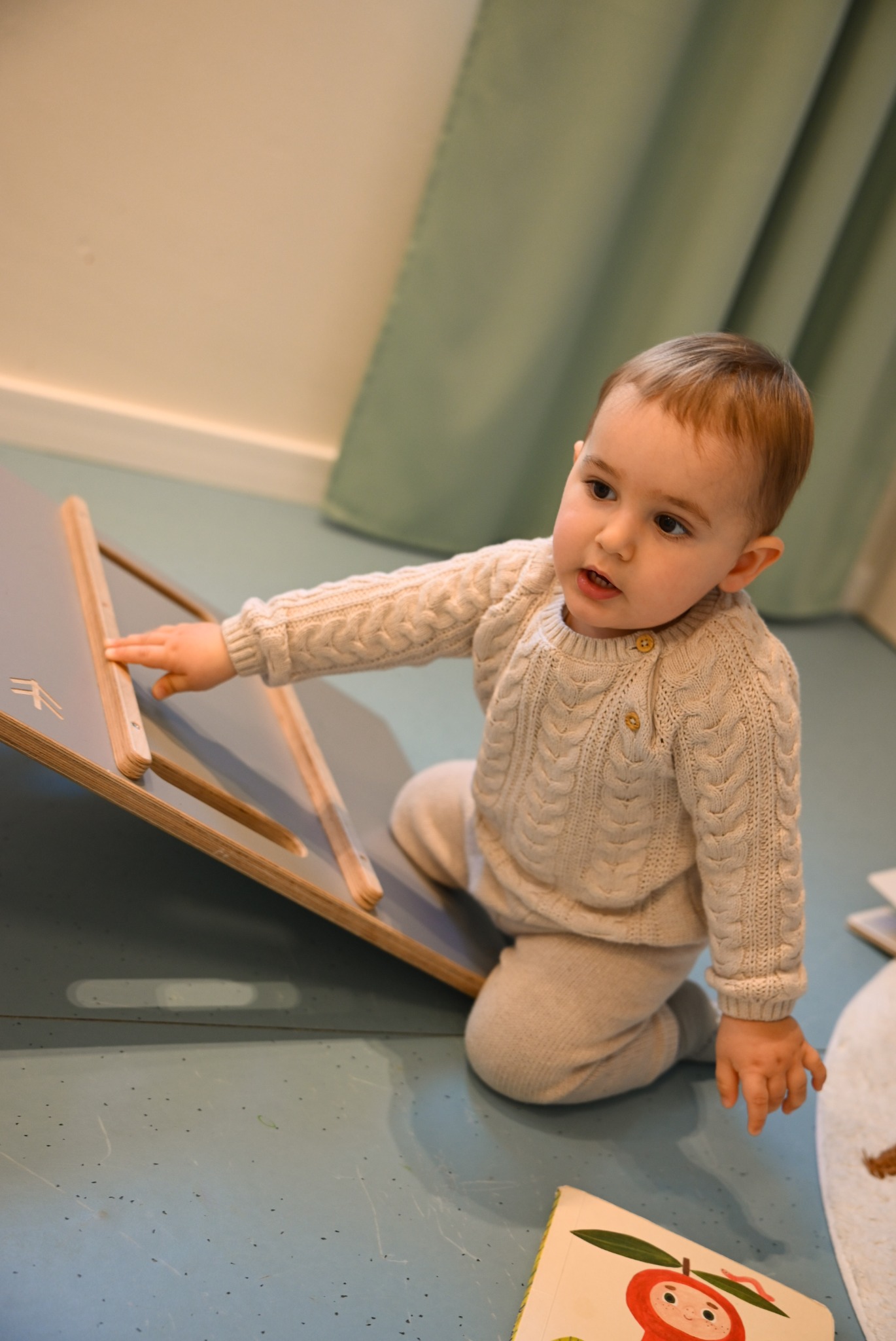 Malutki chłopiec w beżowym swetrze i spodniach bawi się na podłodze pochylnią z drewnianymi szczebelkami, obok leży książeczka z ilustracją jabłka.