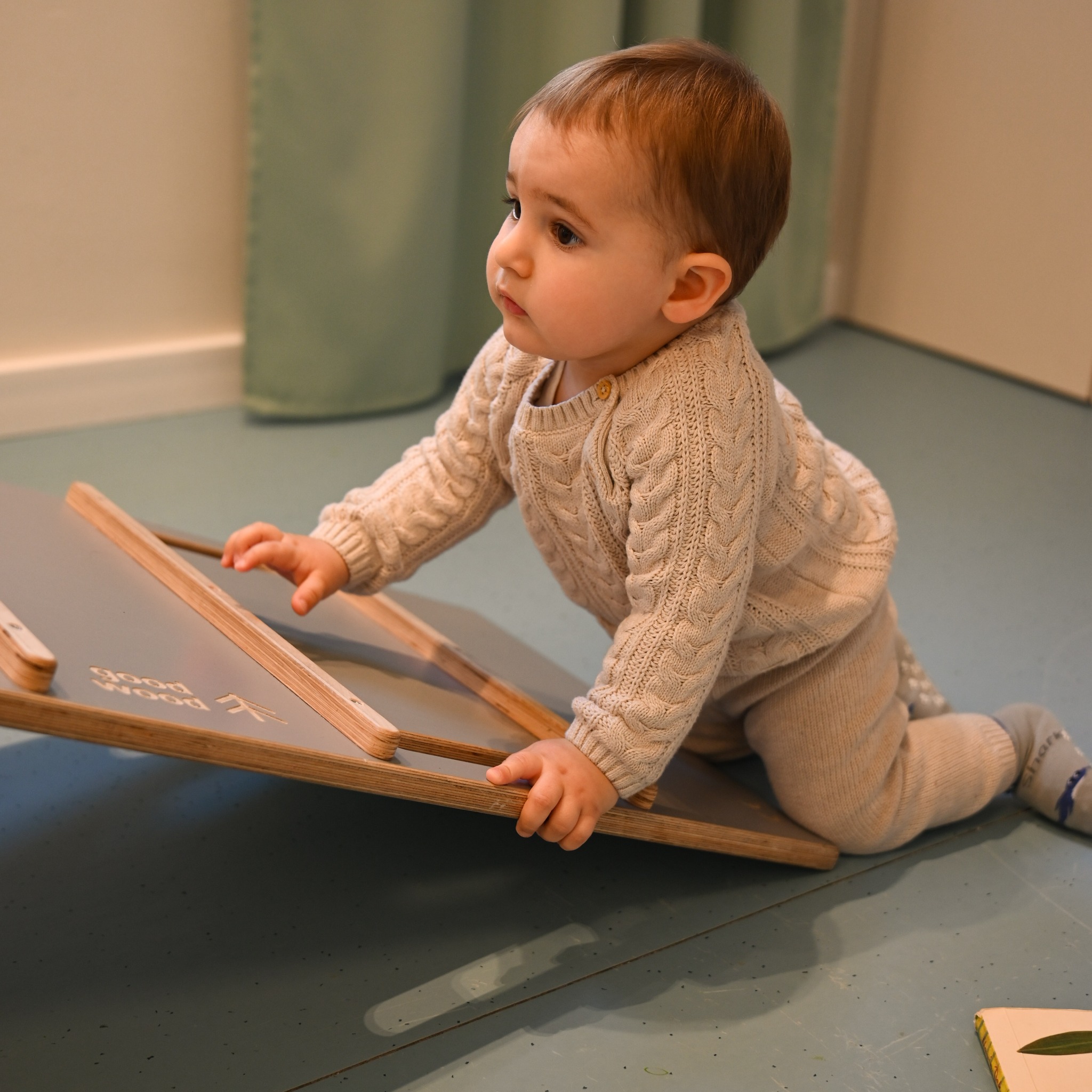 Uroczy niemowlak w beżowym sweterku i spodniach raczkuje po pochylonej drewnianej desce z napisem 'Good Wood', spoglądając w lewo.