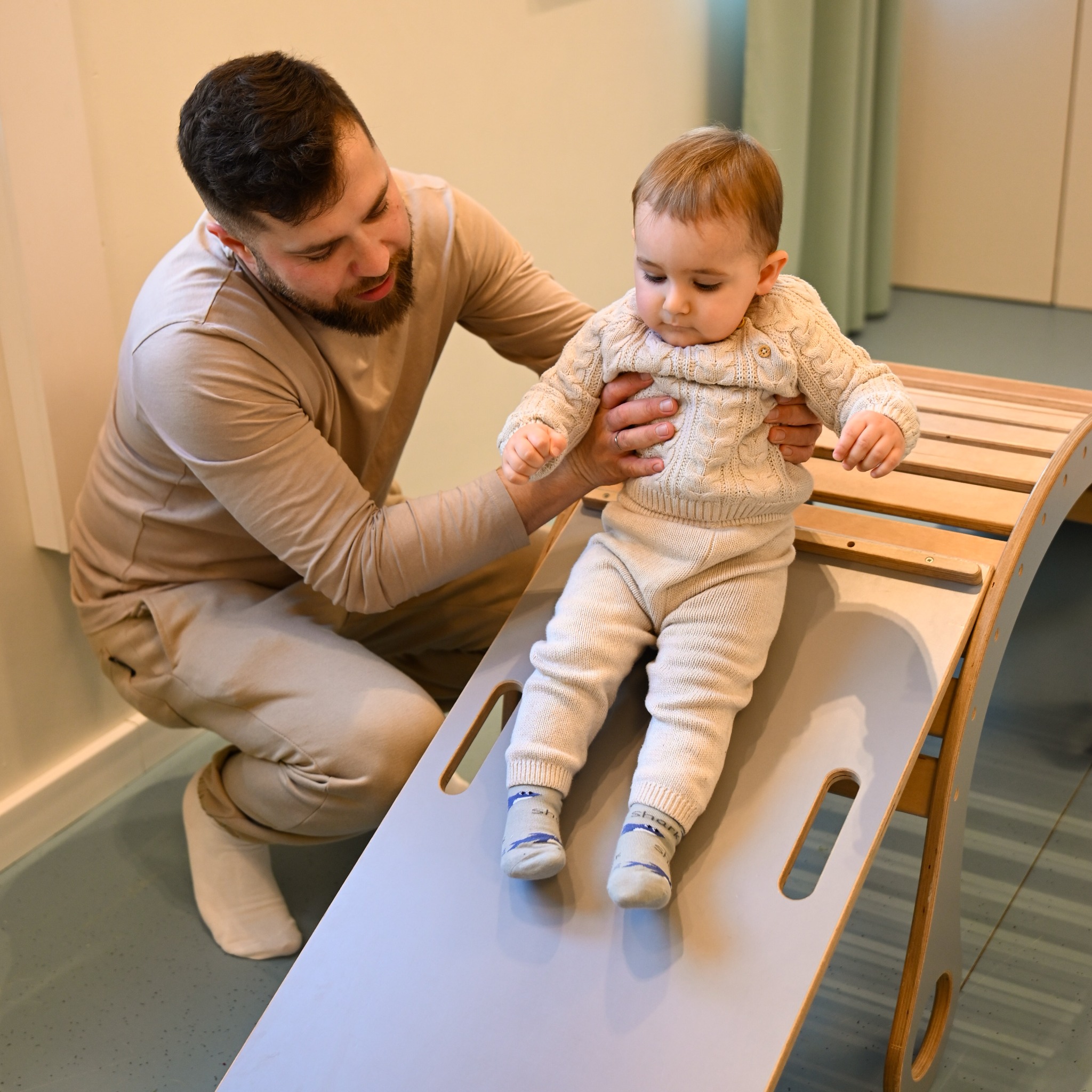 Ojciec asekuruje małe dziecko w beżowym, dzierganym ubranku, podczas zabawy na drewnianej zjeżdżalni w jasnym wnętrzu.