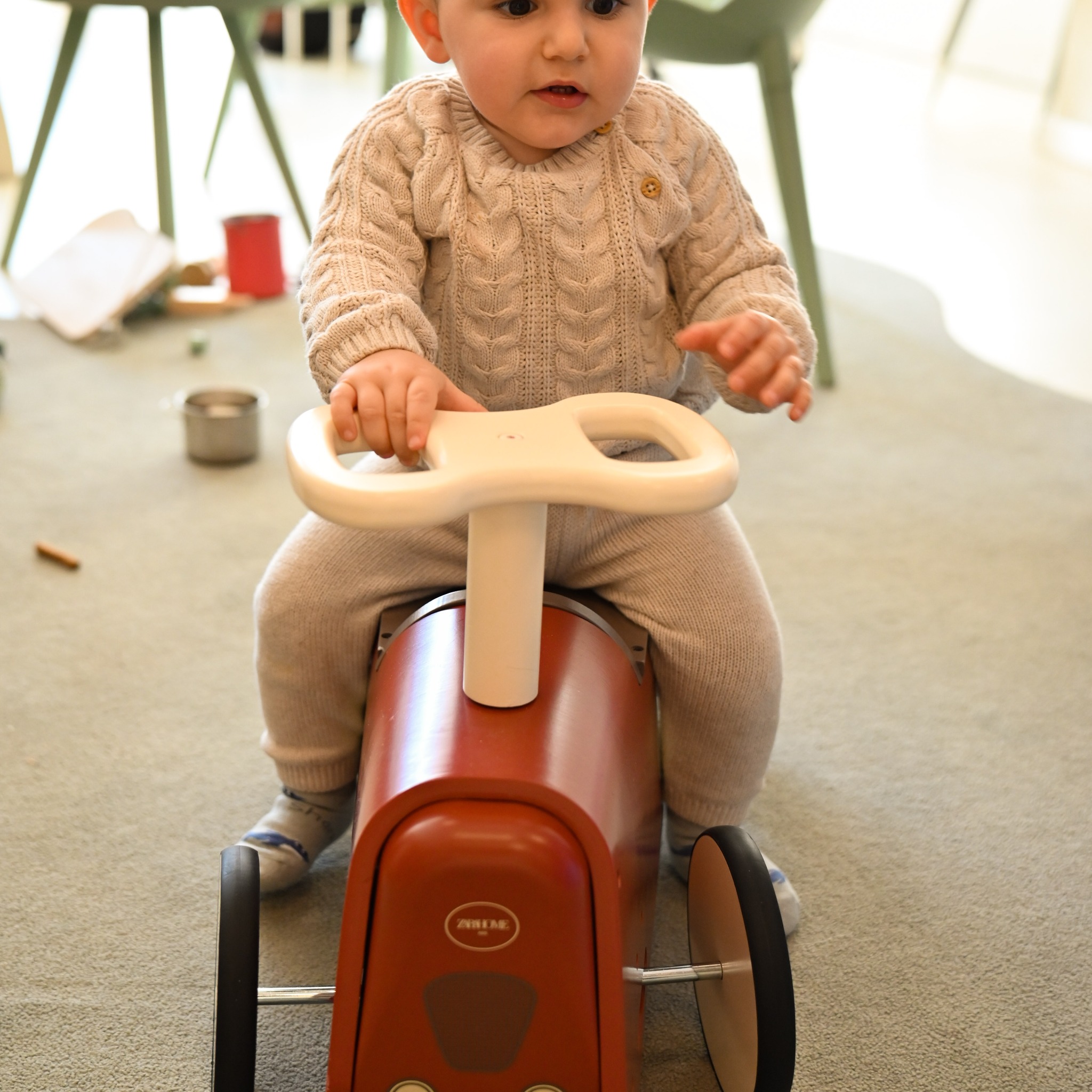 Mały chłopiec w beżowym swetrze siedzi na czerwonym, drewnianym jeździku z białym kierownicą, na dywanie w tle widać zabawki i meble.
