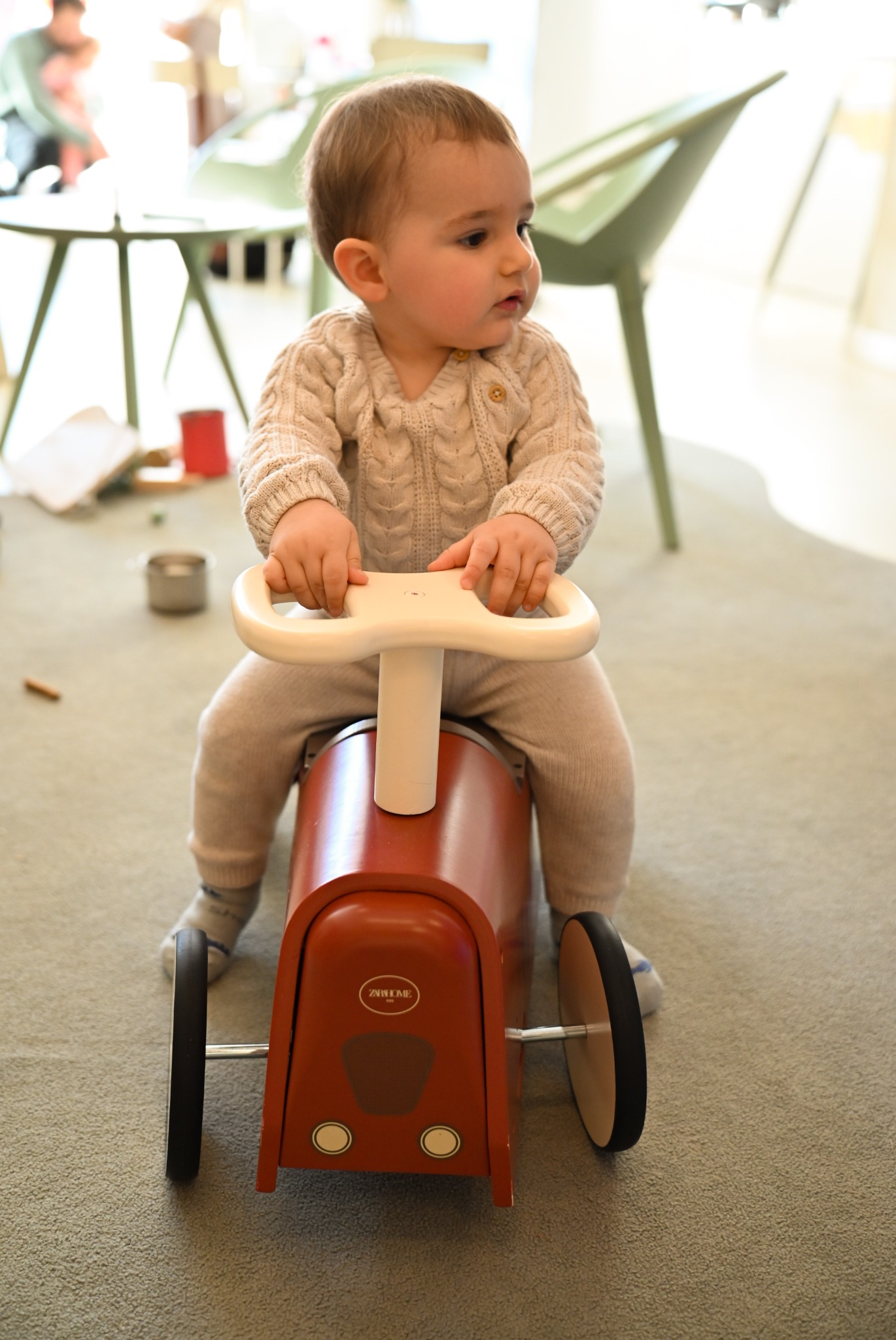 Mały chłopiec w beżowym swetrze i spodniach siedzi na czerwonym jeździku z białym kierownicą w jasnym wnętrzu.