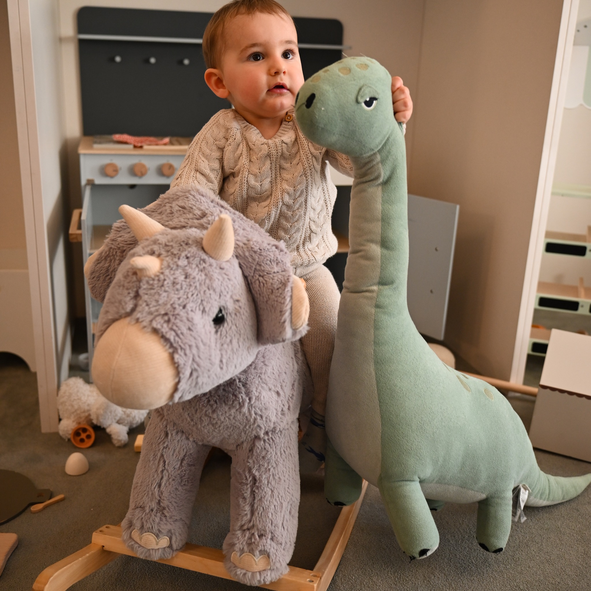 Chłopiec w kremowym swetrze i spodniach bawi się pluszowymi dinozaurami: triceratopsem na drewnianym biegunie i długoszyim brachiozaurem, w tle zabawkowy kuchenny zestaw.